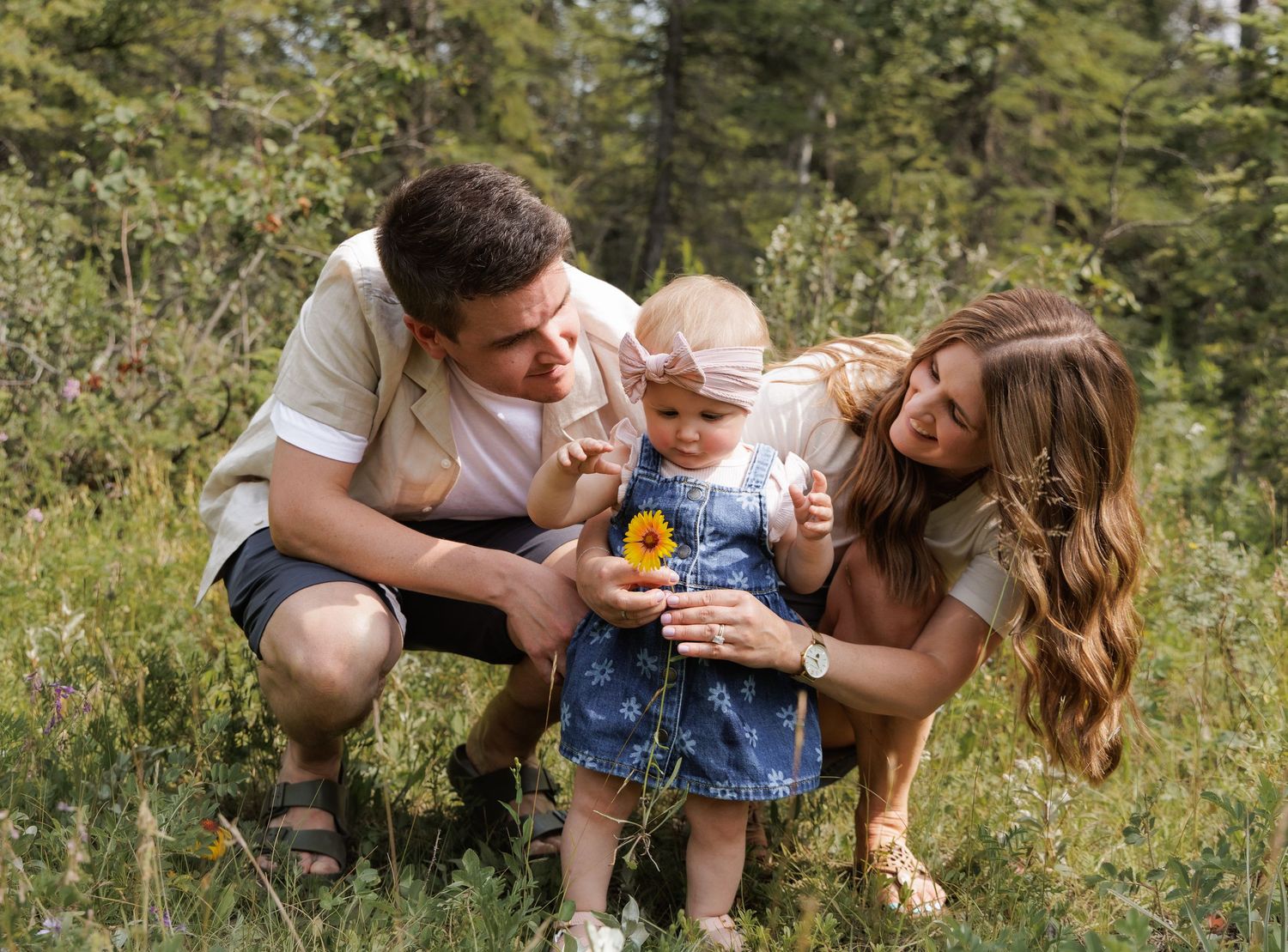 A young family with a baby girl picking wildflowers in a lush green forest meadow during a sunny day.