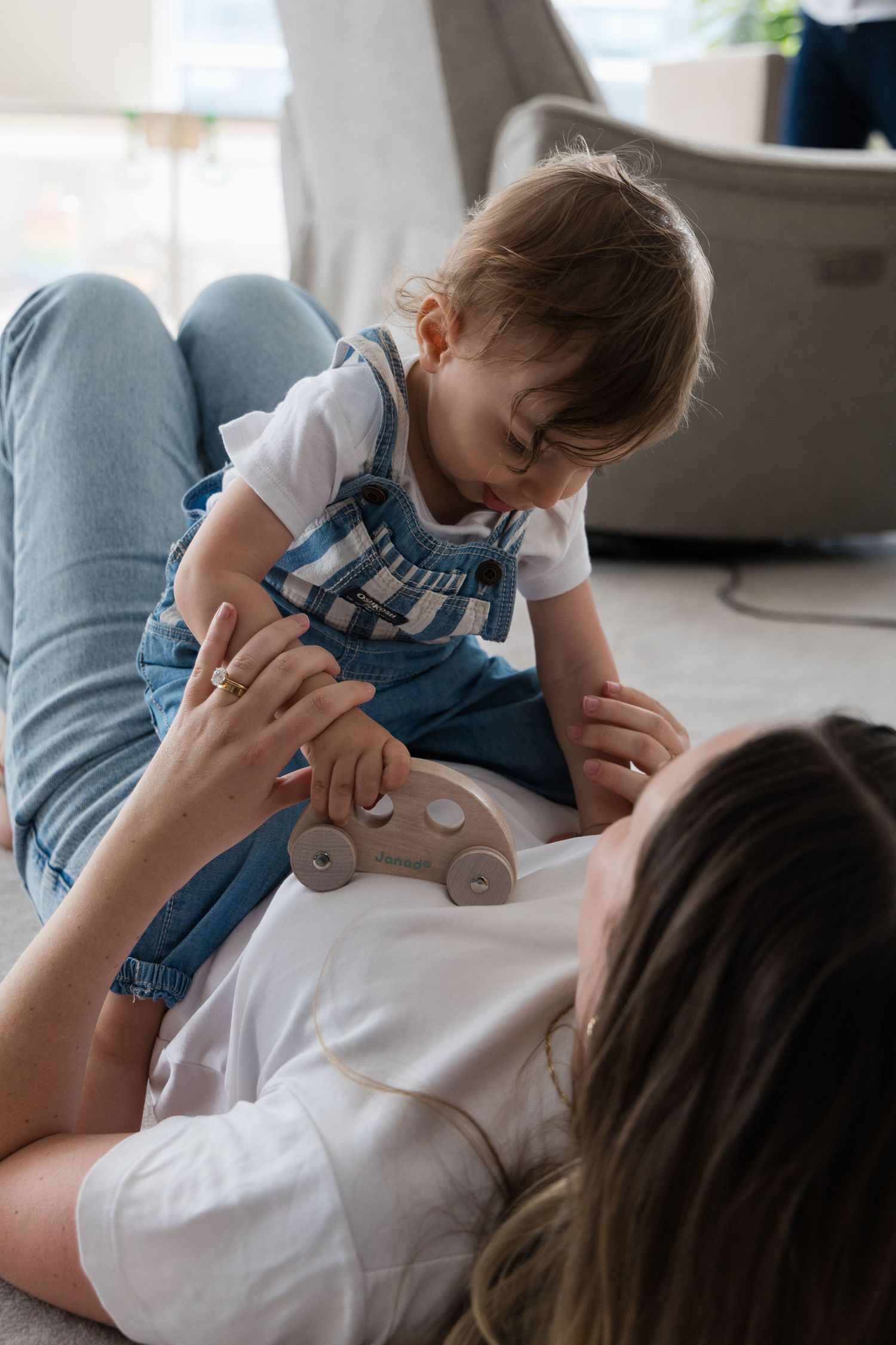 Mother lying on floor playing with toddler holding a wooden toy car.