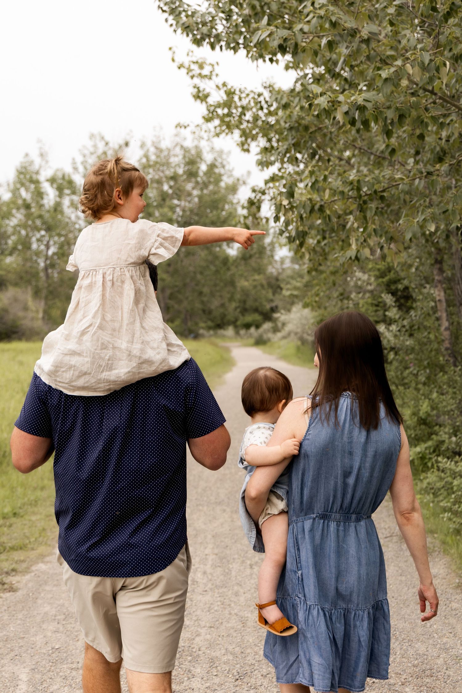 Family walking on a dirt path, father carrying child on shoulders, mother holding baby, surrounded by green trees.