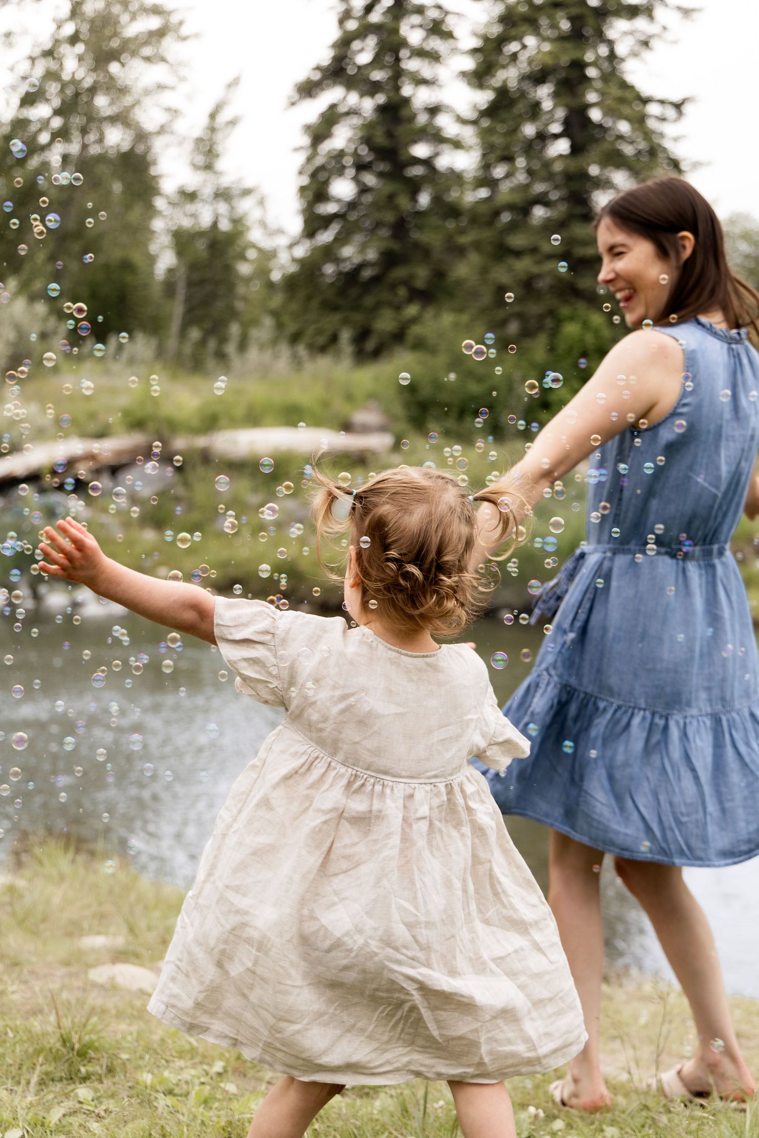 A laughing mother in blue dress plays with her toddler daughter among floating bubbles near a pond.
