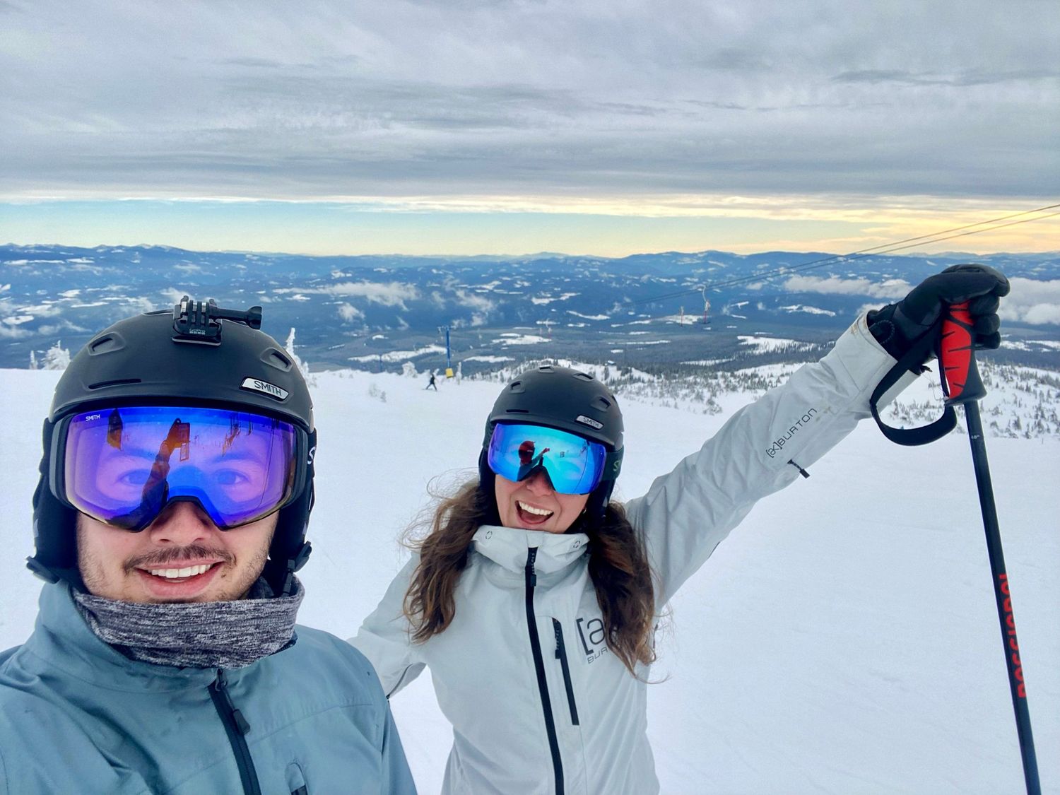 Two skiers smiling in helmets and goggles taking a selfie on a snowy mountain summit.