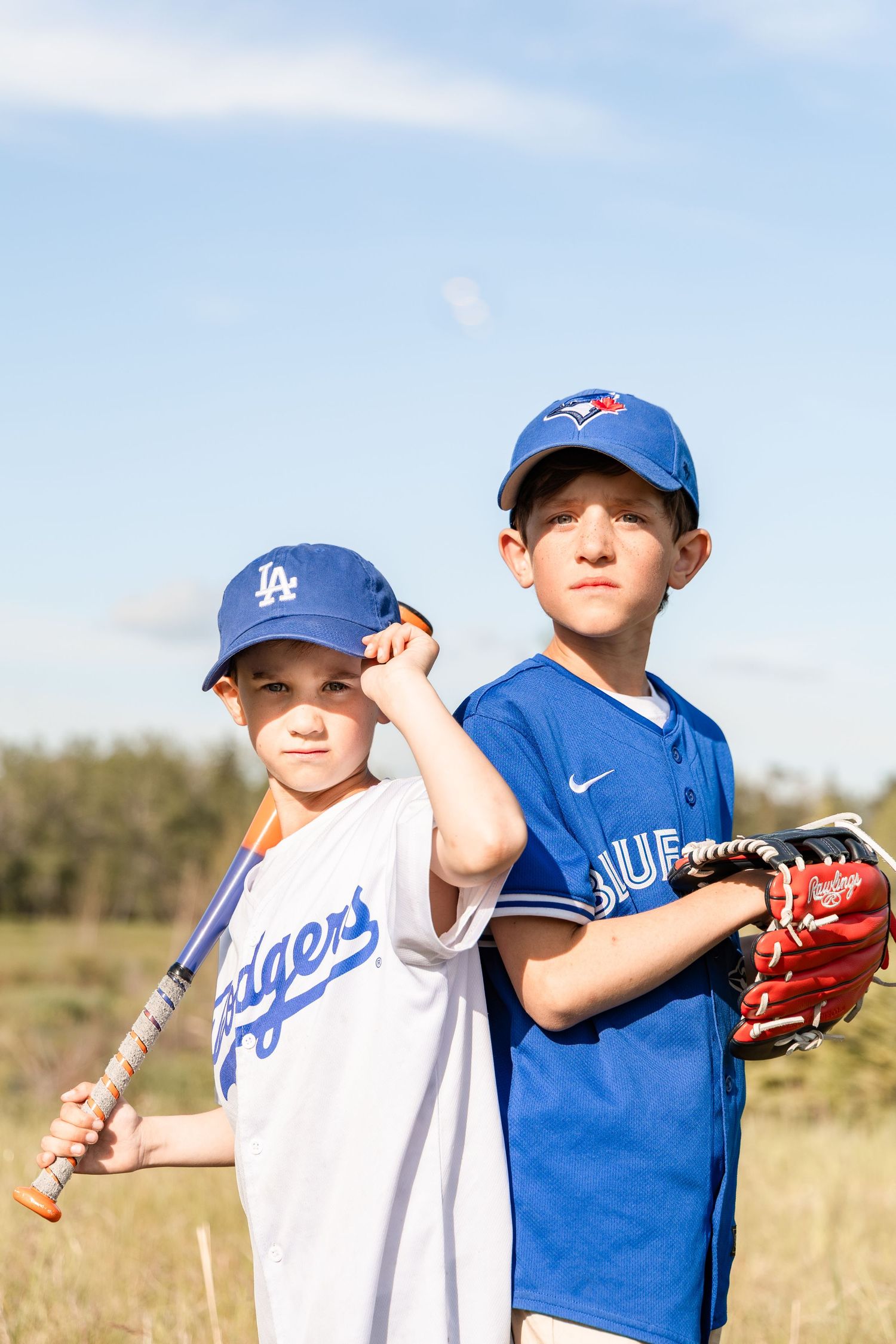 Two boys in baseball uniforms pose outdoors, one in a Dodgers jersey and one in a Blue Jays jersey.