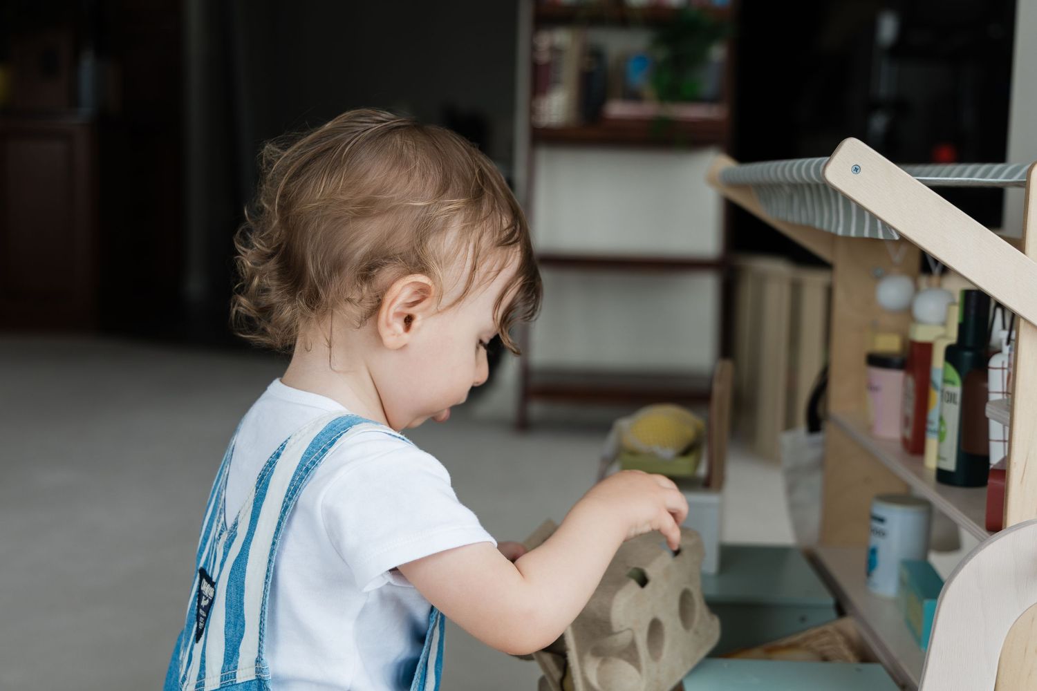 Side view of toddler in striped overalls exploring a wooden play kitchen set.