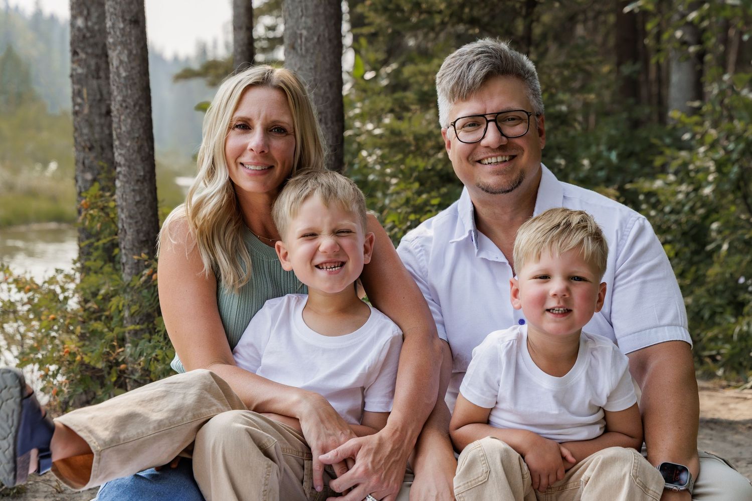 Happy family of four smiling outdoors in a forest setting, parents with two young blonde boys.