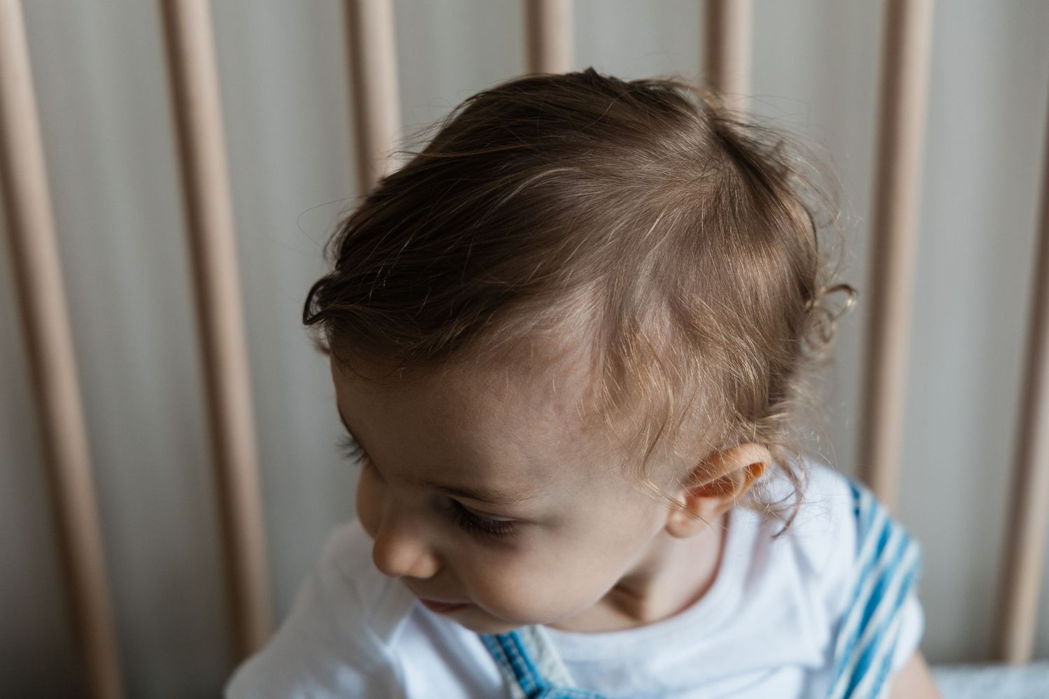Close-up of a baby with curly hair standing in a wooden crib looking down.