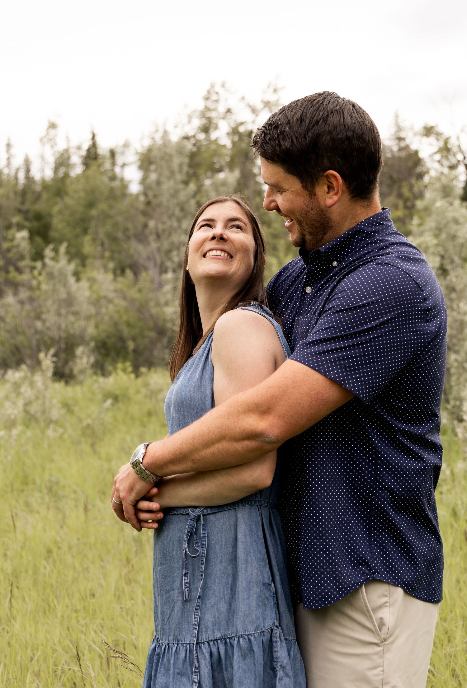 A smiling couple embraces in a lush green meadow, the woman looking up at the man lovingly.