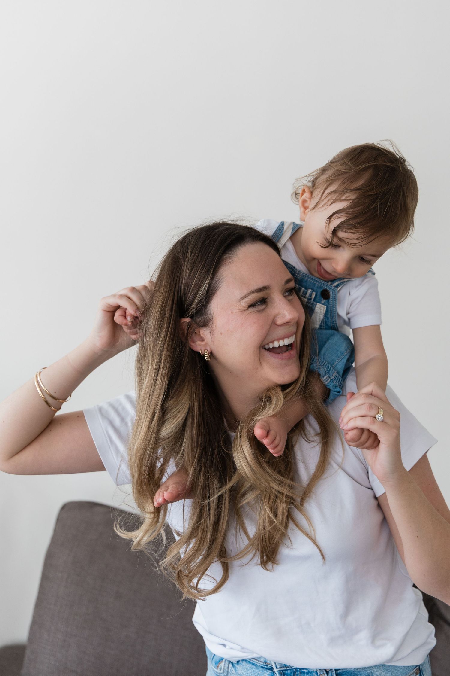 Laughing mother in white tee holds her giggling toddler on her back in a bright living room.