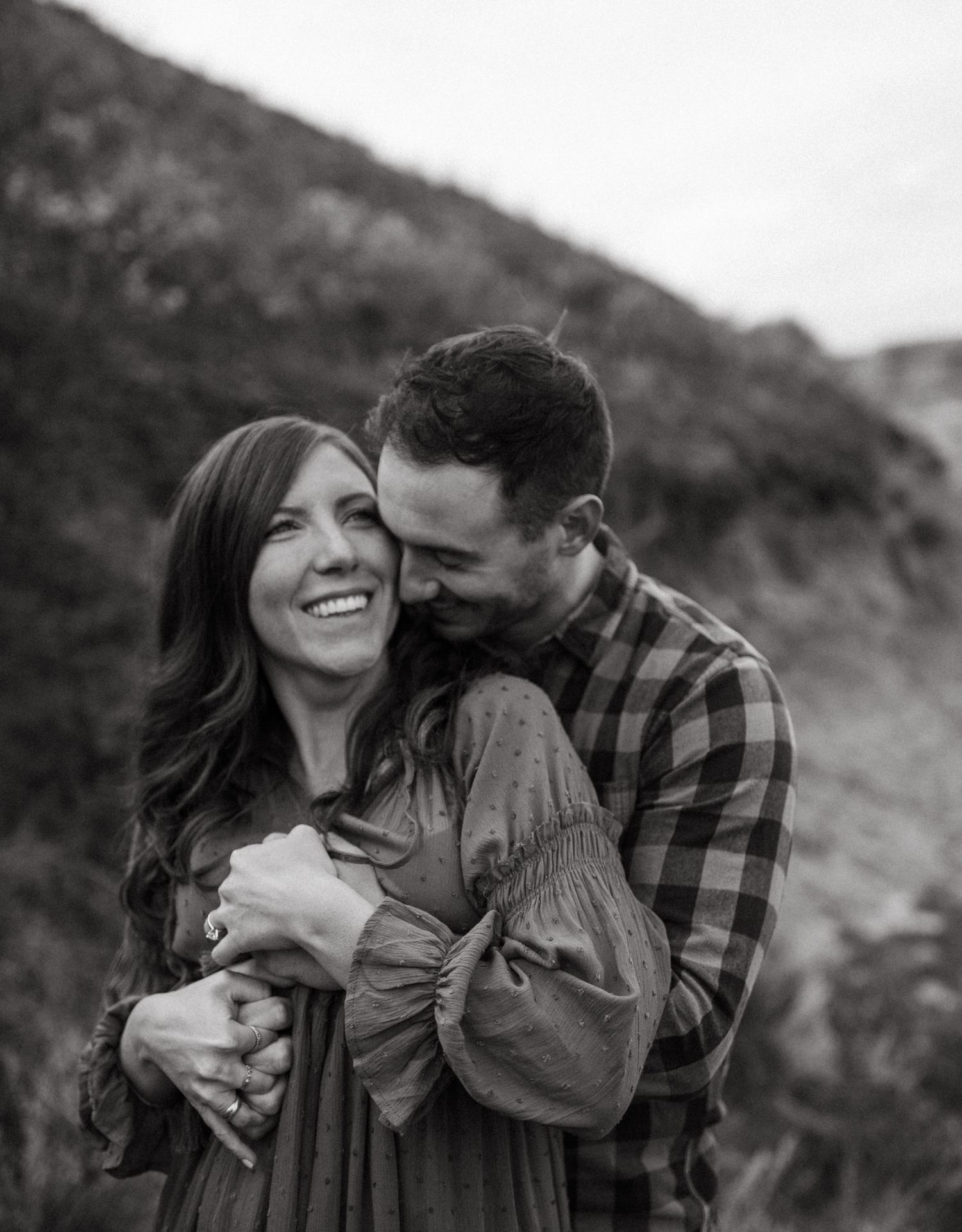 Black and white romantic photo of a couple embracing and laughing outdoors near a hillside.