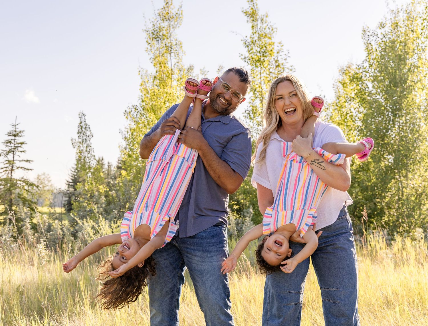 Happy family of four playing outdoors in a sunny meadow, parents swinging two girls upside down while laughing.