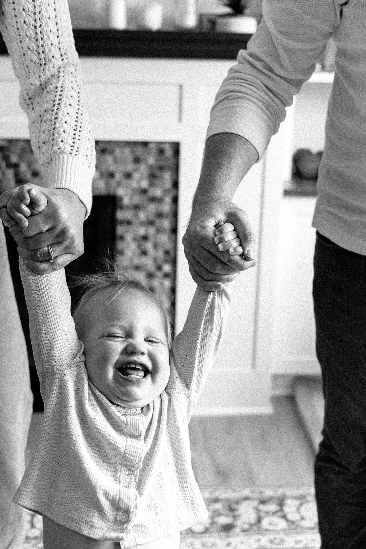 Black and white photo of joyful baby laughing while parents hold hands in kitchen.