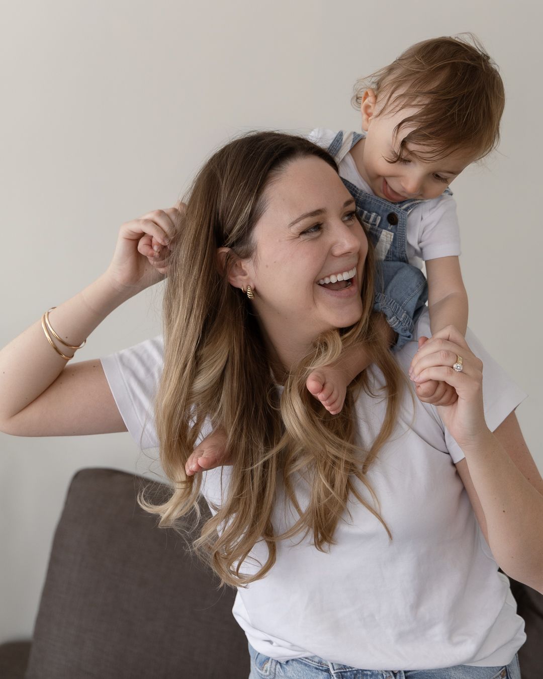 A laughing woman in a white tee carries a happy toddler on her shoulders indoors.