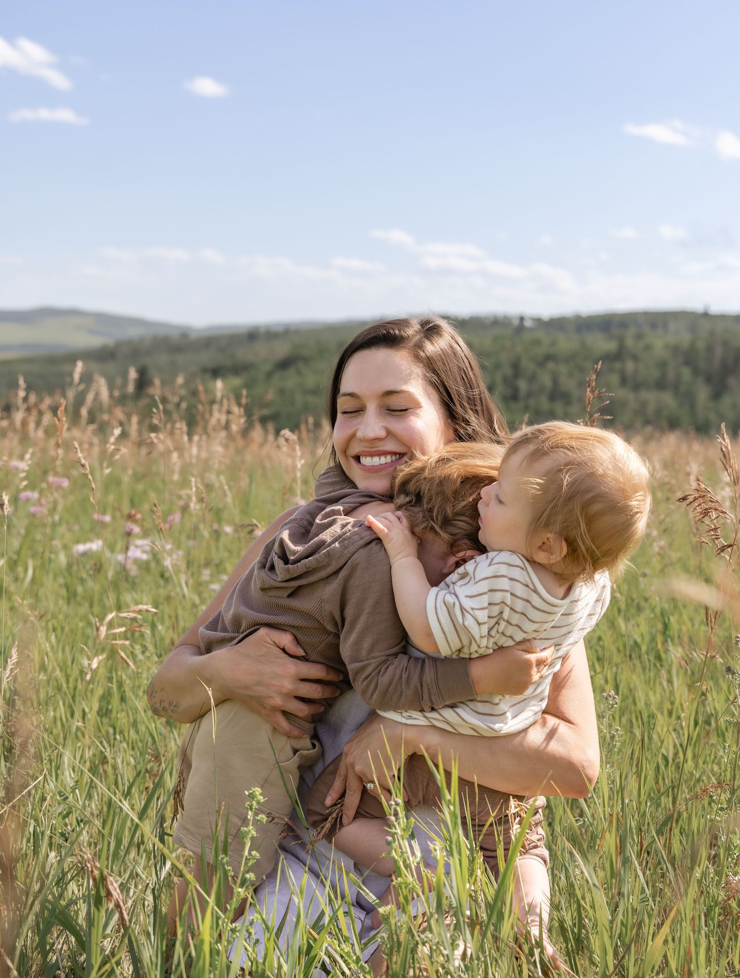 A smiling woman hugs a toddler in a sunny green meadow with rolling hills in the background.