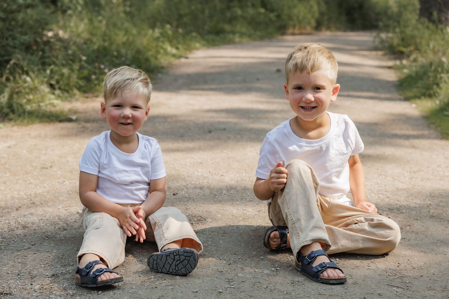 Two young blonde boys in white t-shirts and khaki pants sitting on a dirt path, smiling outdoors.