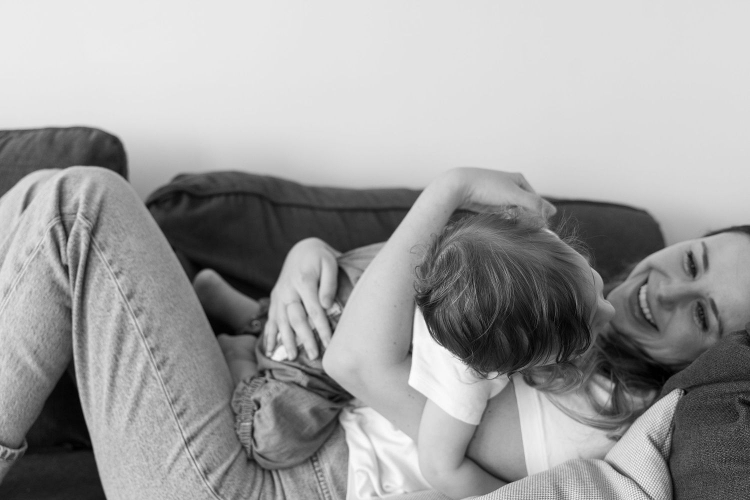 Black and white photo of mother and toddler cuddling and laughing together on a gray sofa.