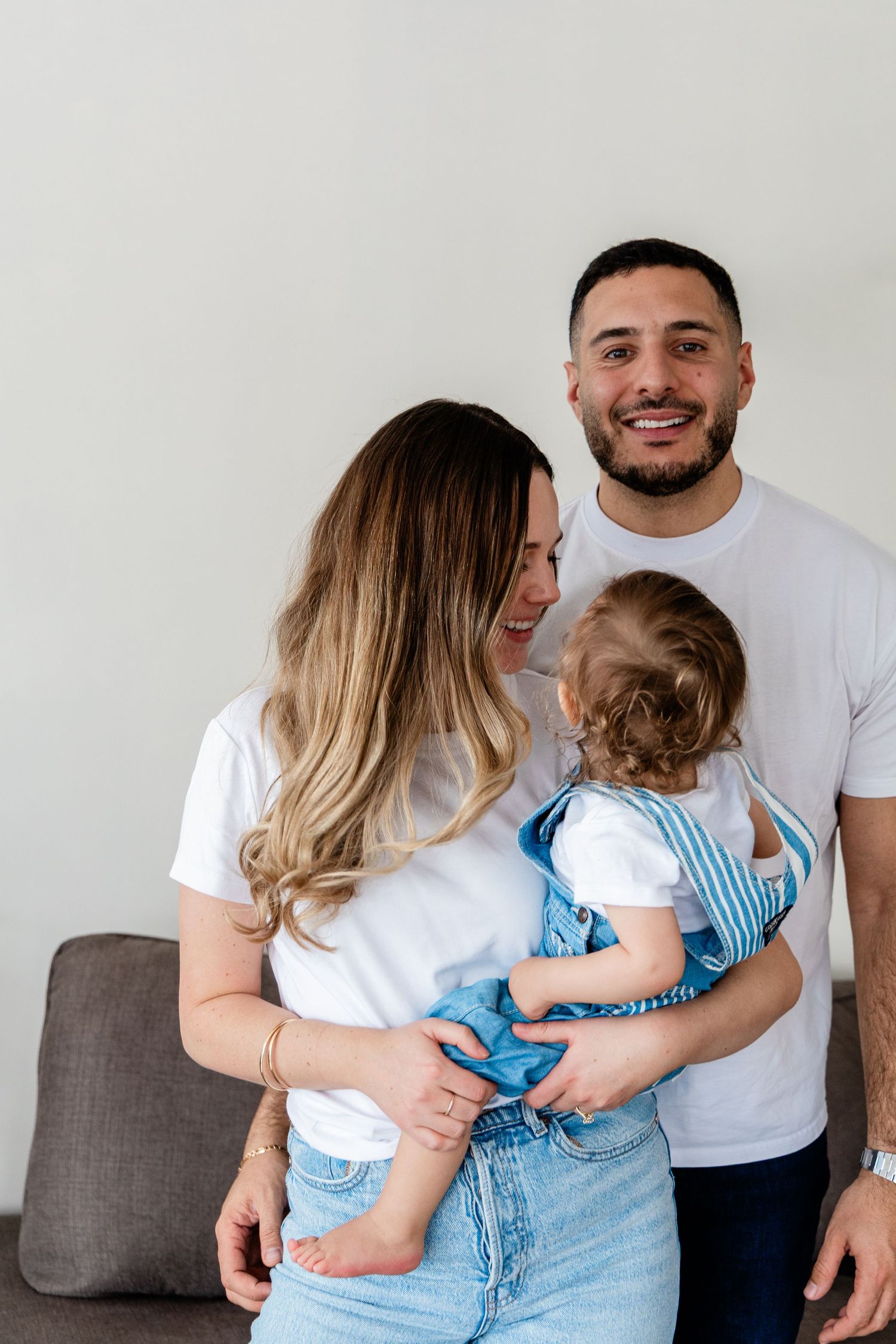 Happy family with mother holding baby in blue overalls while father smiles warmly.