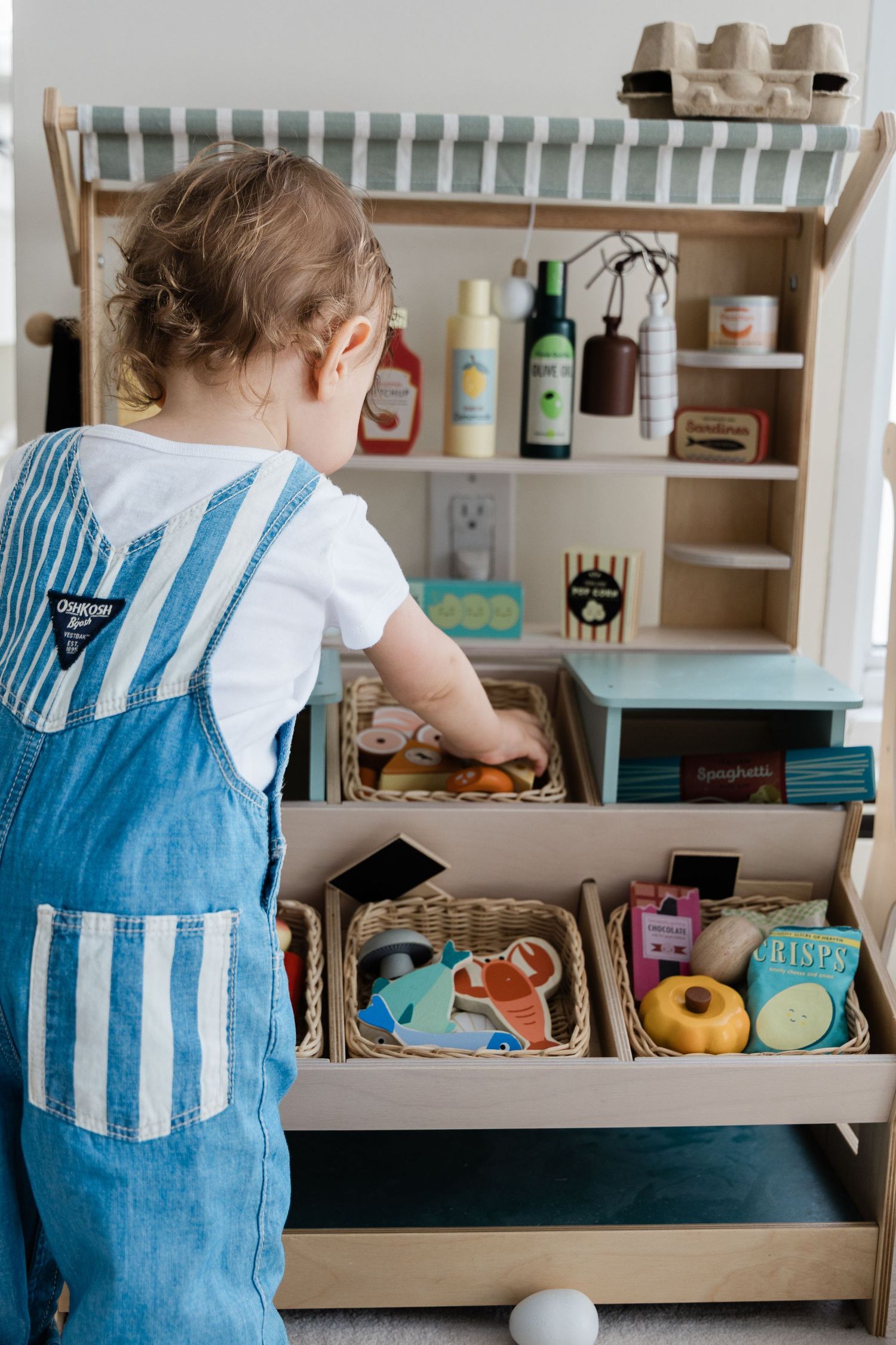 Toddler in denim overalls playing with a wooden toy market filled with colorful pretend food and goods.