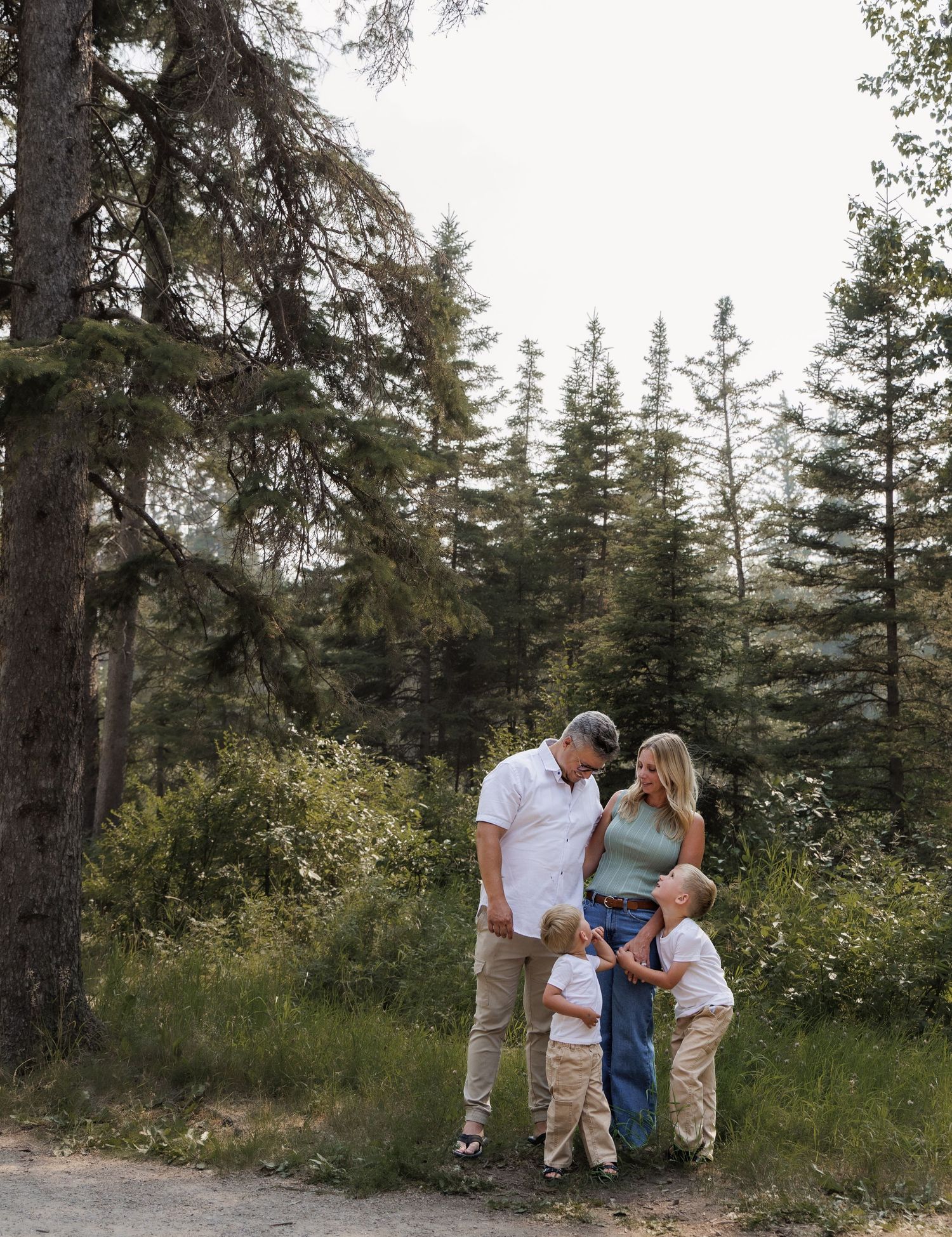Family of four with two toddlers sharing a joyful moment on a forest trail surrounded by tall pine trees.