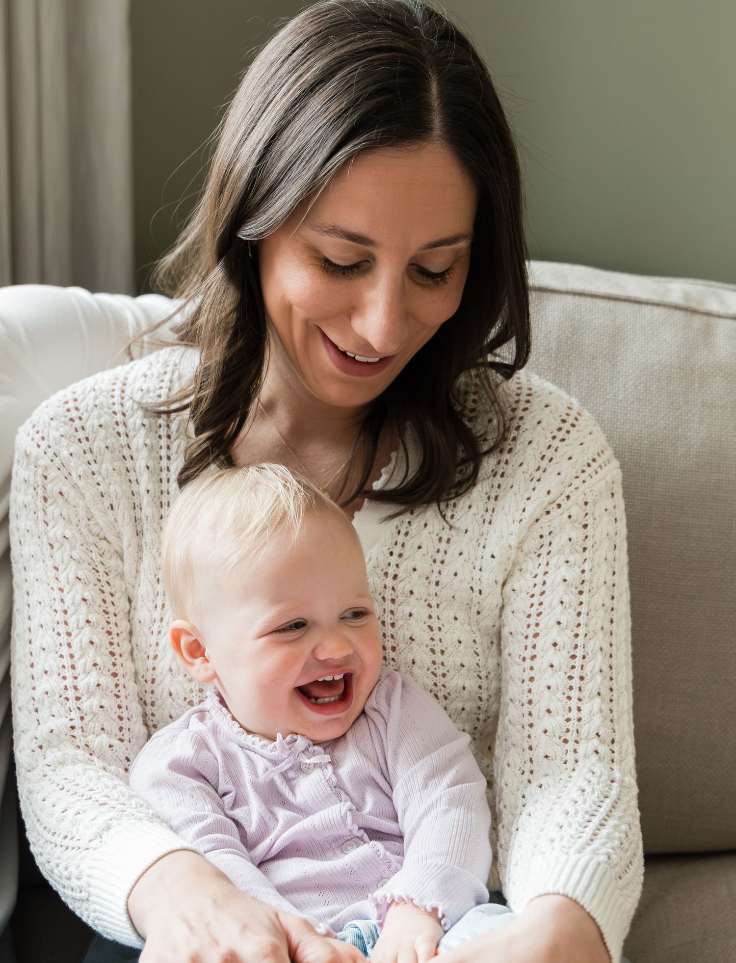 Smiling mother in white sweater holding her laughing blonde baby girl on couch.