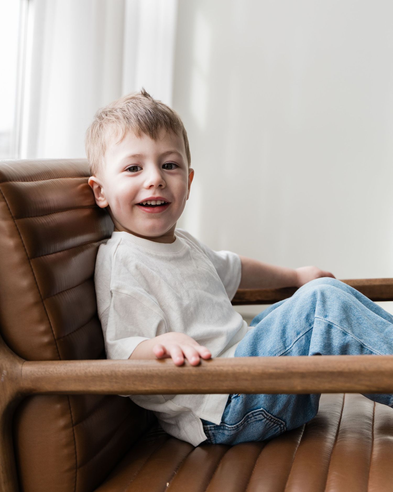 Happy young blonde boy smiling and sitting relaxed in a brown leather armchair near a window.