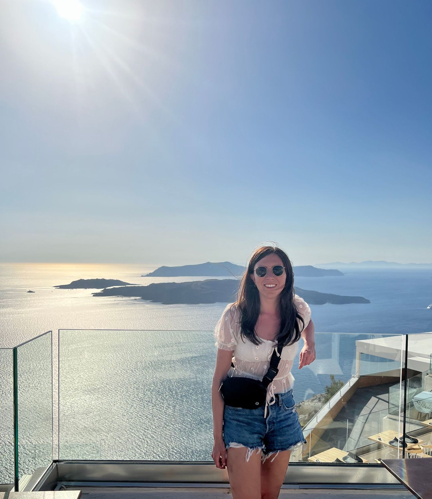 A woman in denim shorts smiles on a sunny overlook with a stunning Santorini caldera view behind her.