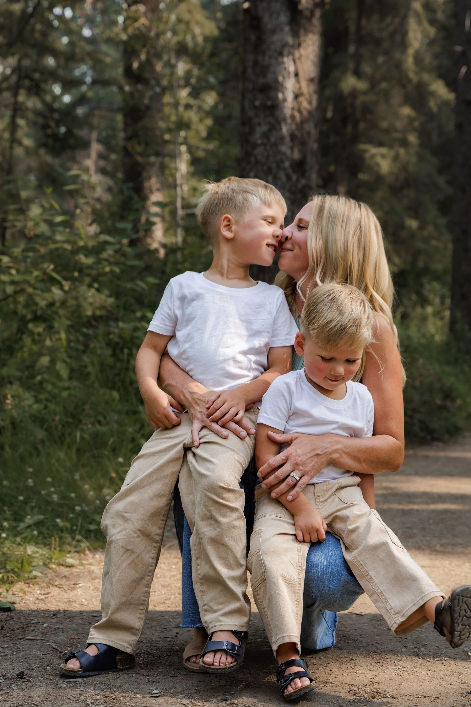 A blonde mother kisses her young son while holding two boys in white shirts and beige pants on a forest path.
