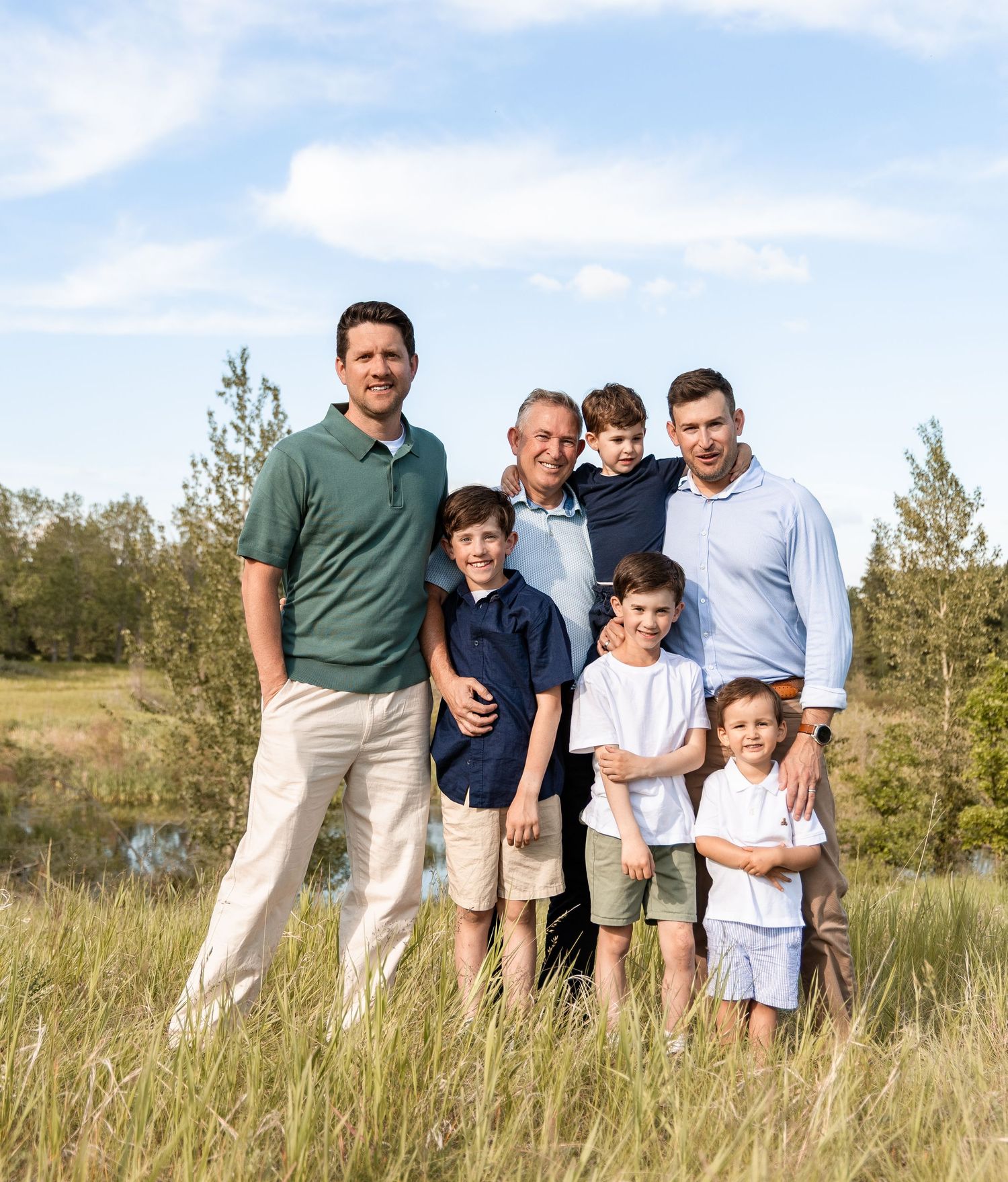 Three generations of men and boys pose together smiling in a sunny green field with trees behind.