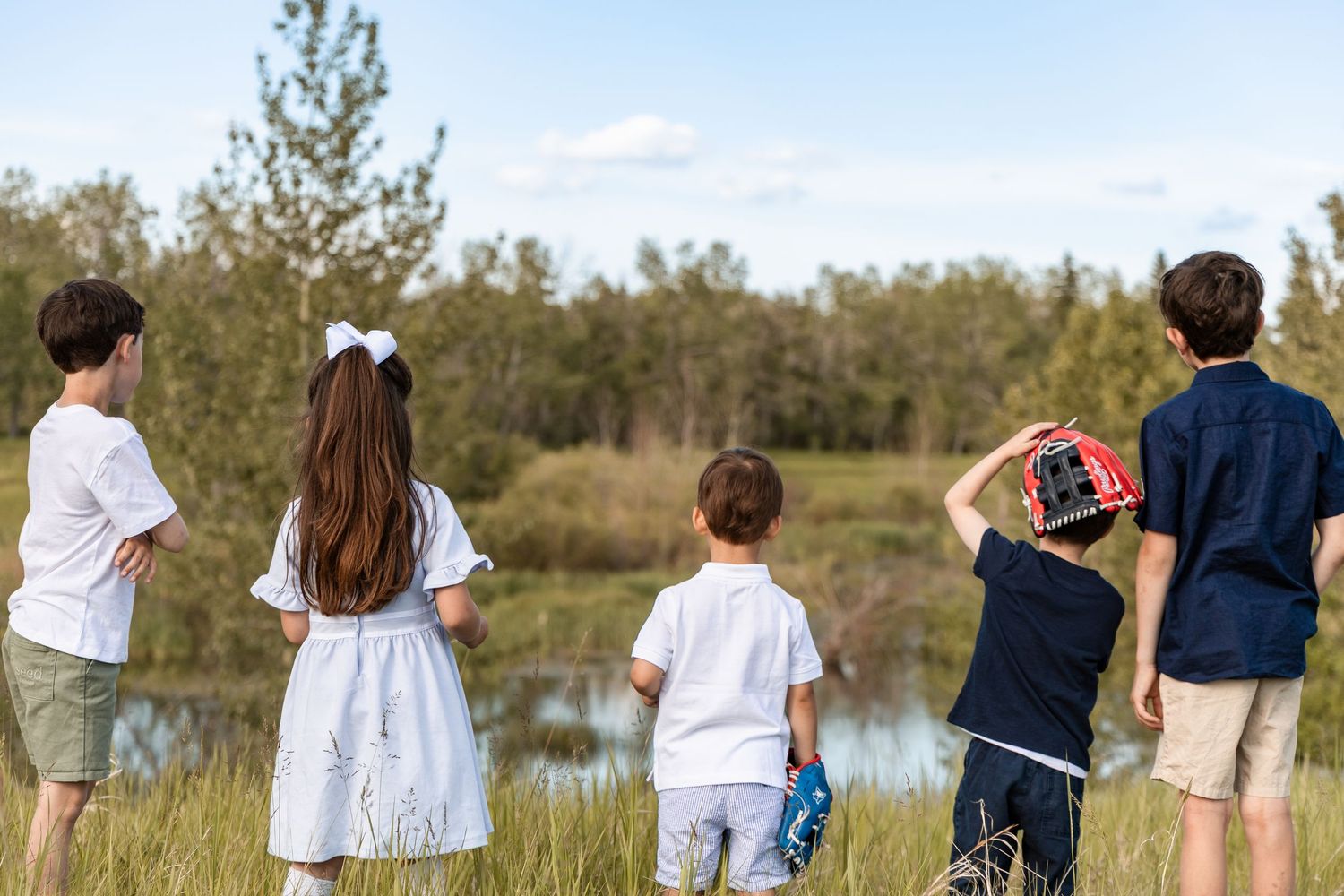 Four children standing outdoors viewing a scenic pond surrounded by trees.
