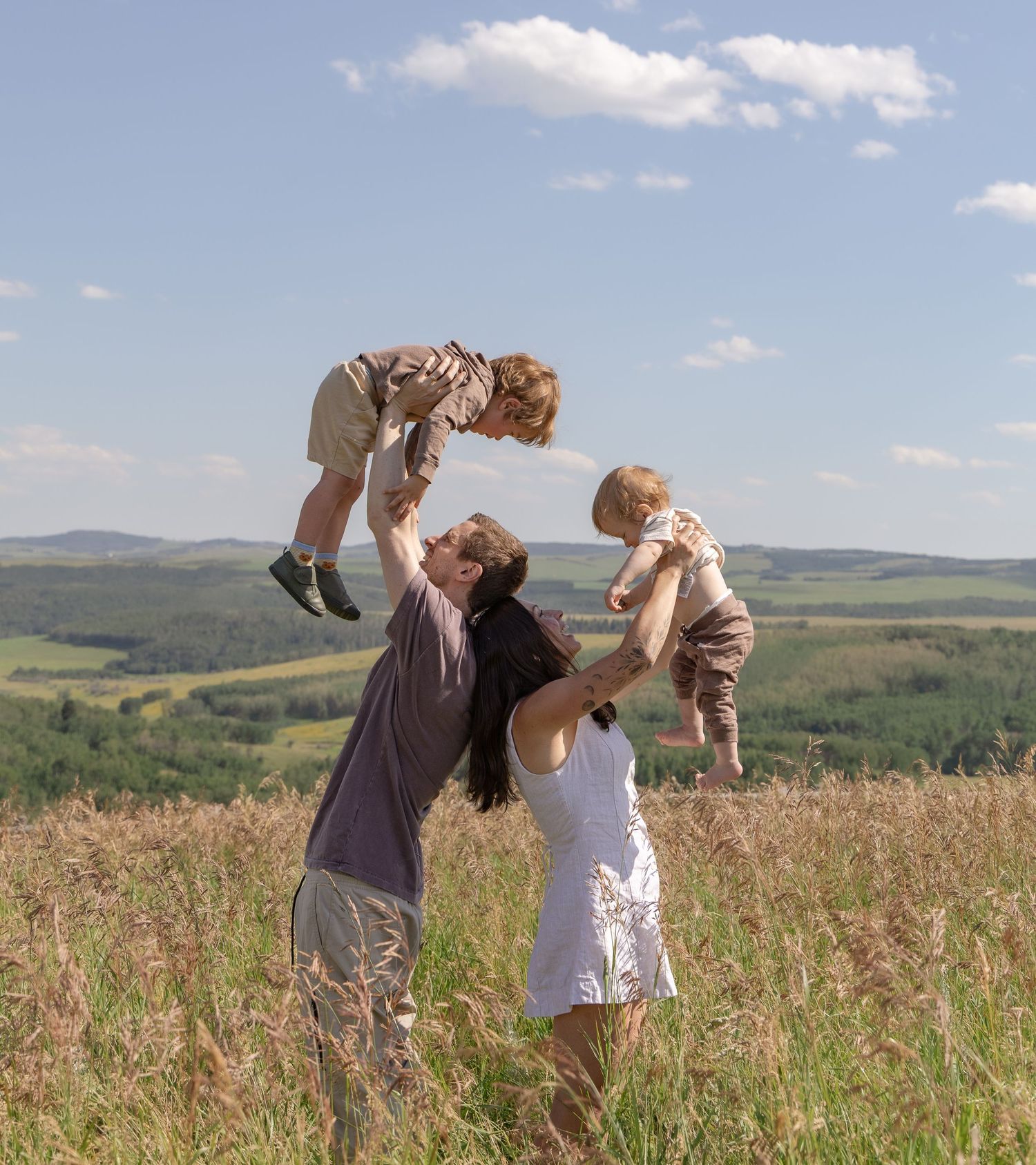 A joyful couple lifts two toddlers in a sunny meadow with rolling hills and blue sky in the background.