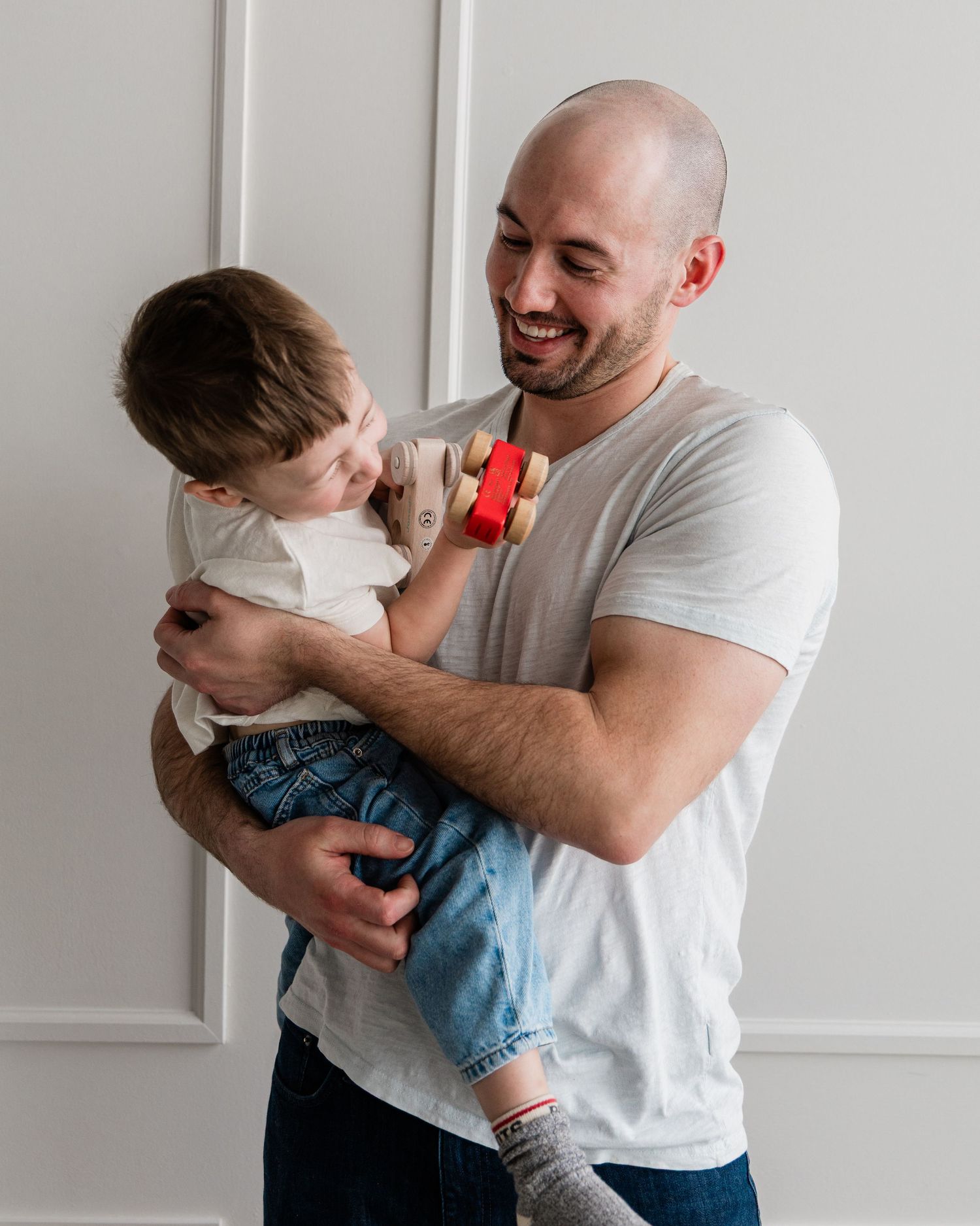 Smiling bald man holding a toddler boy playing with a red toy in a bright white room.