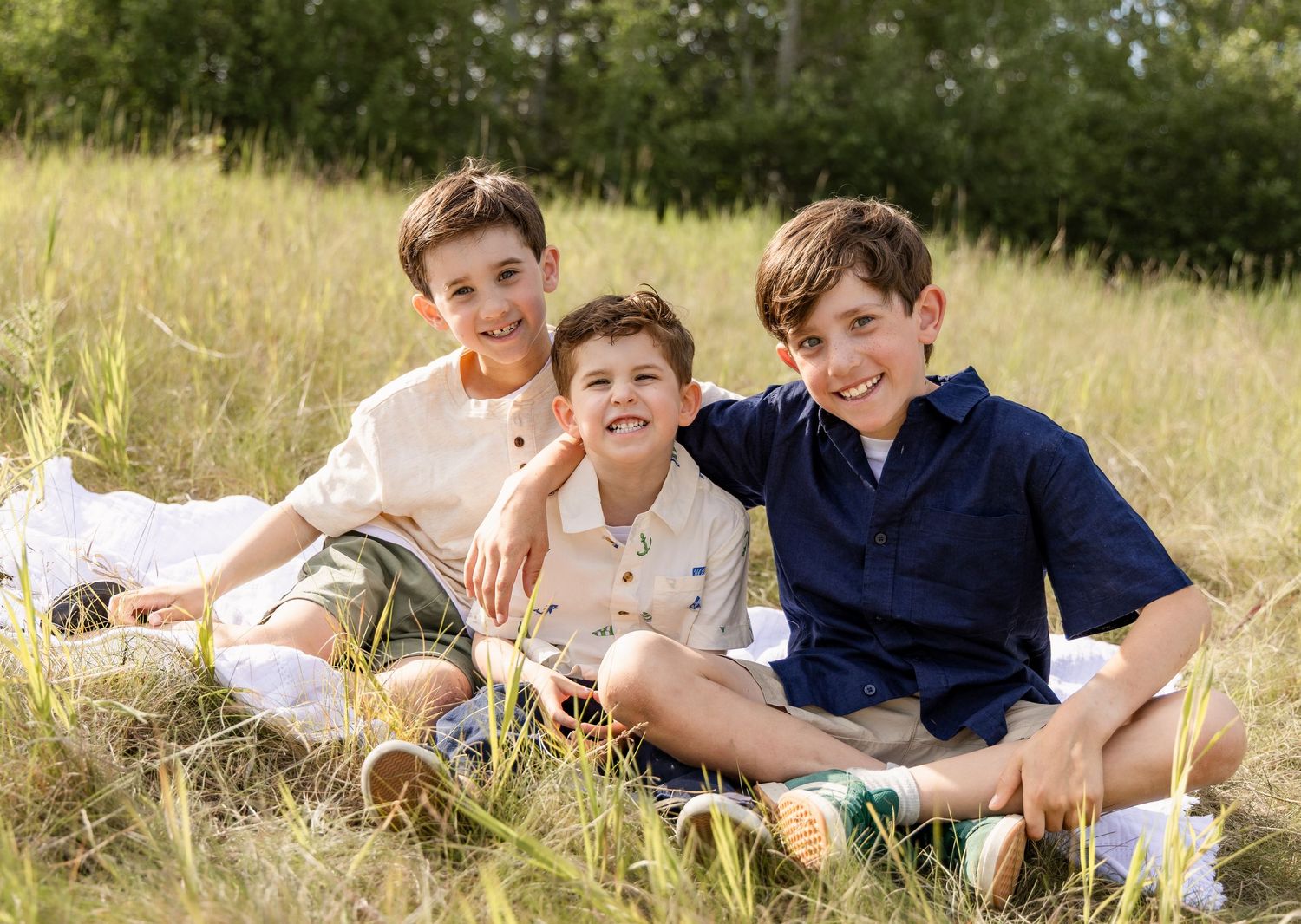 Three smiling brothers sit together in a golden grass field for a warm outdoor family portrait.