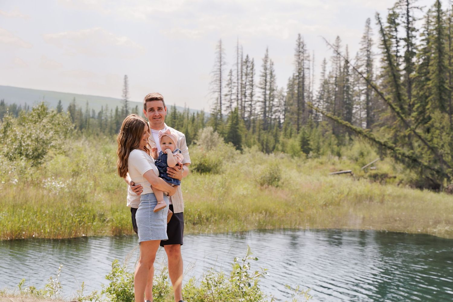 A young couple holding a baby stands by a serene mountain lake surrounded by lush green forest.