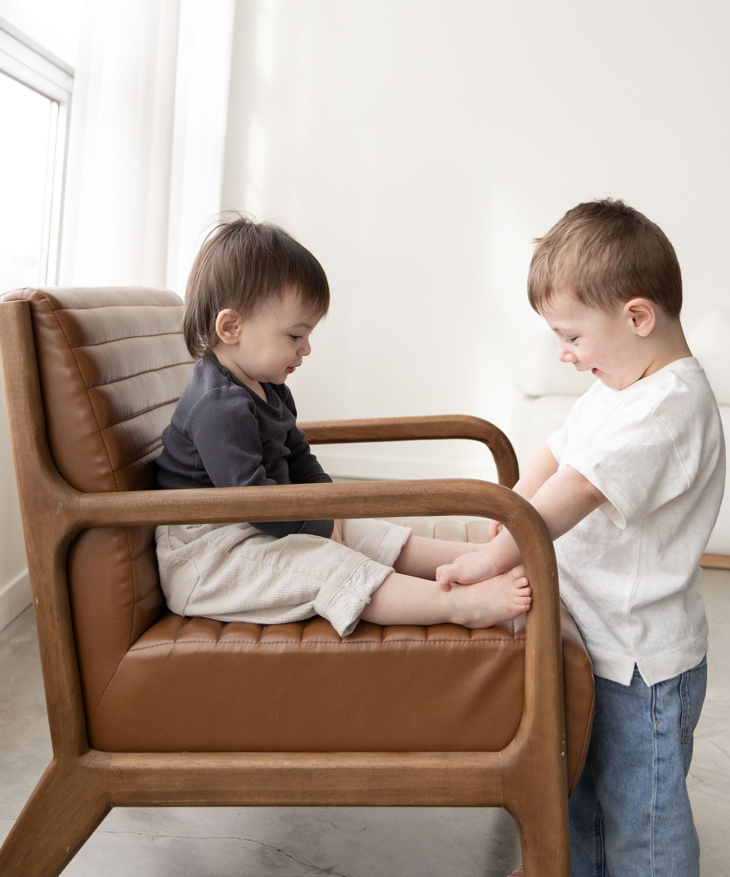 Two young brothers playing together on a brown leather armchair in a bright living room.
