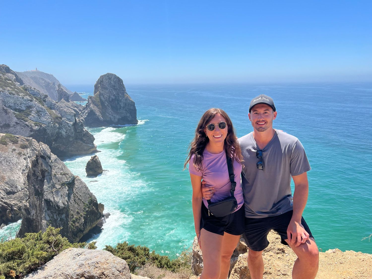 A couple poses on a coastal cliff overlooking turquoise ocean waters and dramatic rock formations.