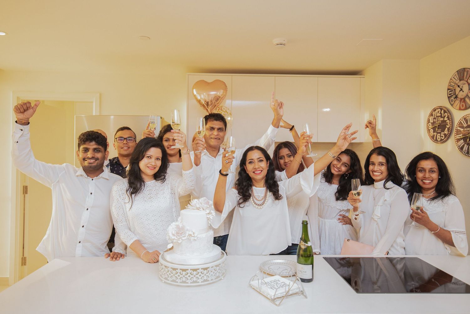 Candid event photography of a large group in white clothing cheering around a celebration cake indoors.