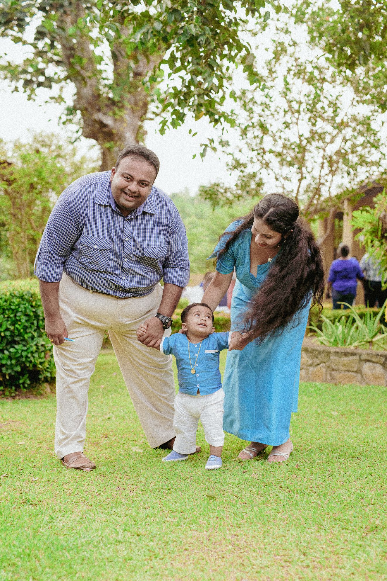 Outdoor family lifestyle session featuring parents and a toddler walking through a bright, green park.