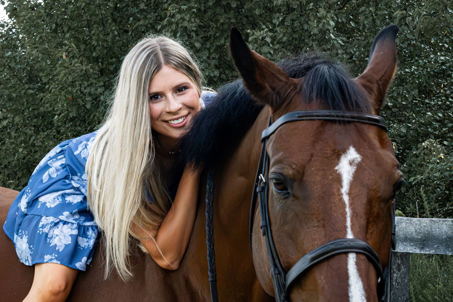 Smiling blonde woman in blue floral top leaning affectionately against a brown horse with a white blaze outdoors.