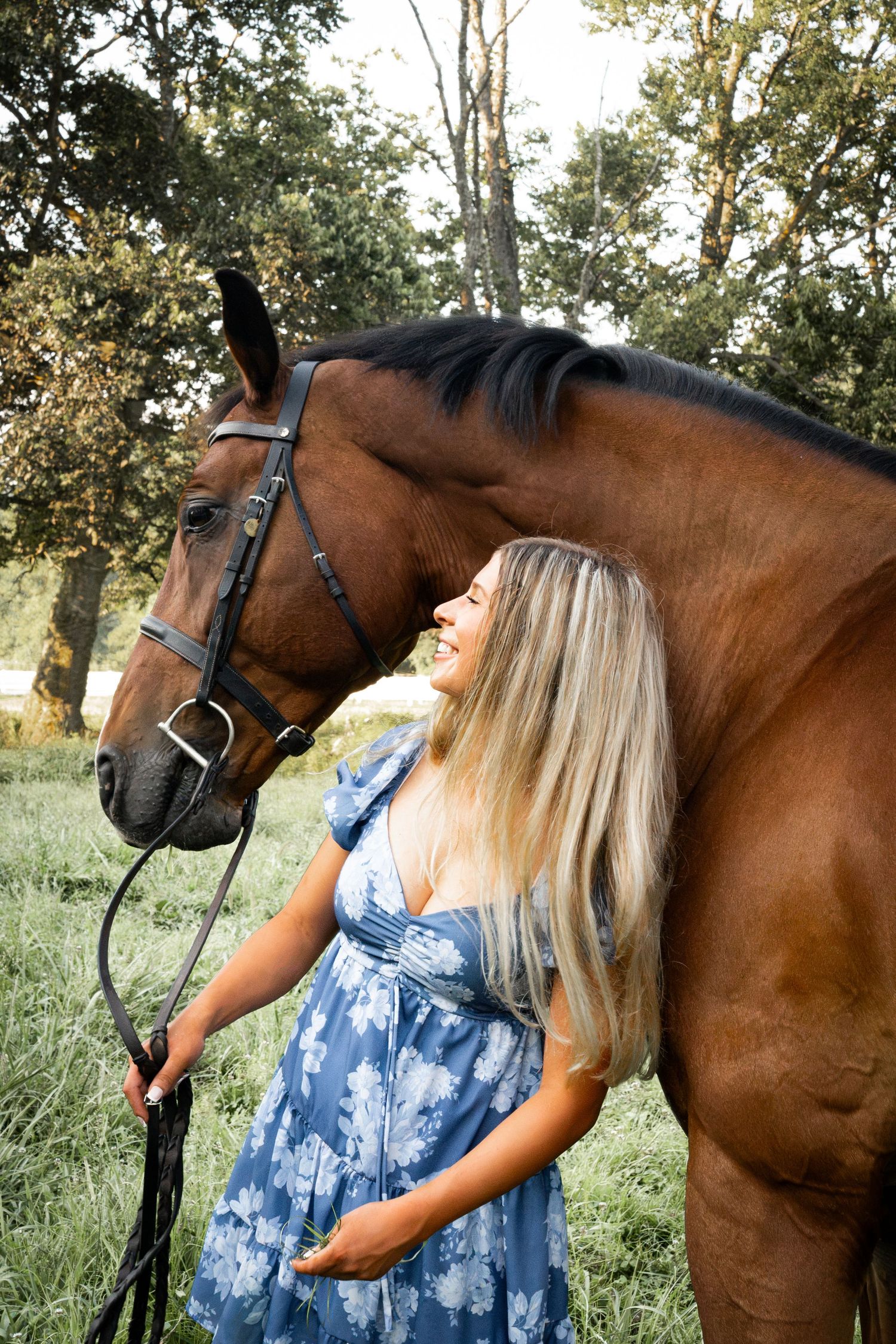 Smiling blonde woman in blue floral dress standing with a bay horse in a lush green outdoor setting.