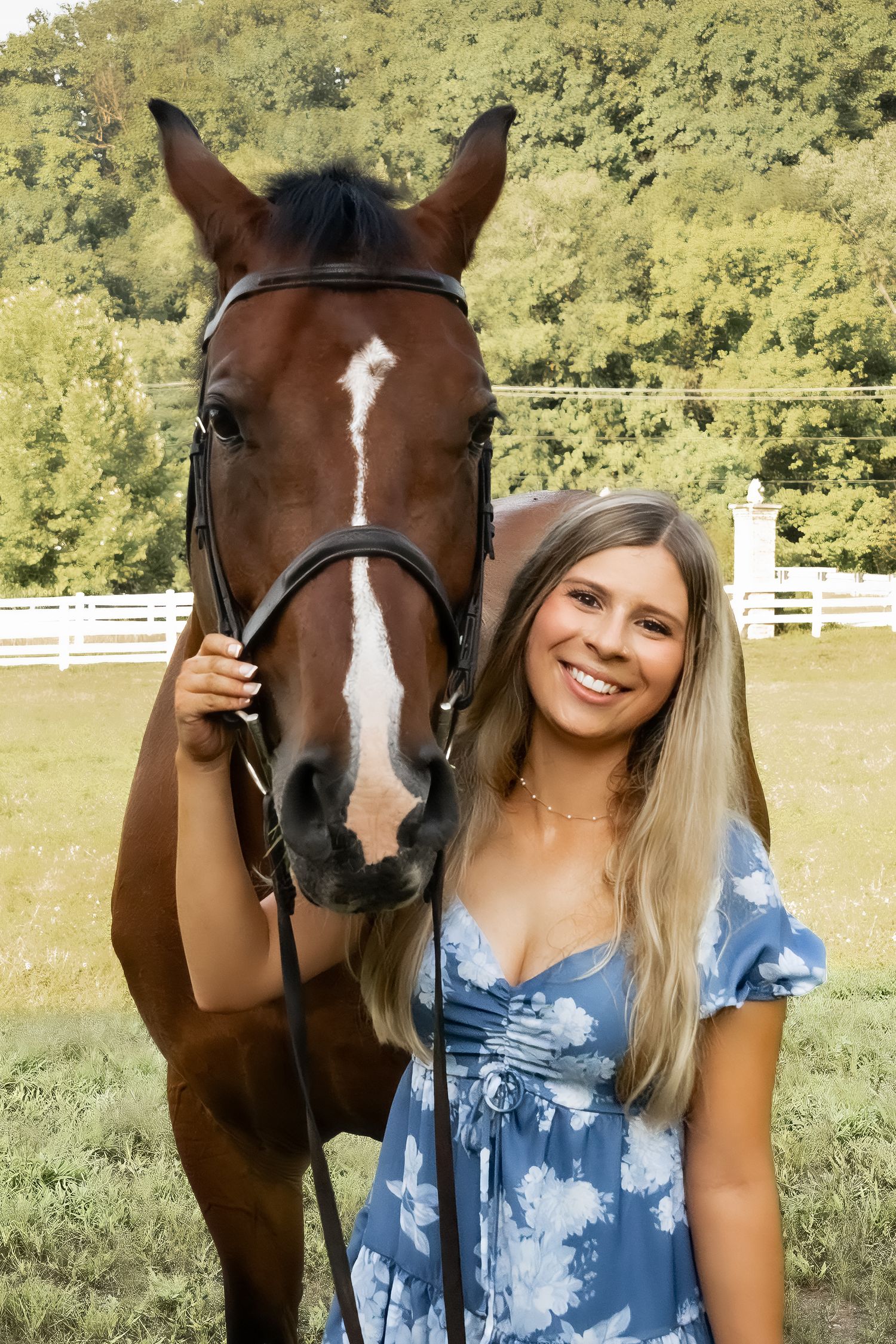 Young woman in blue floral dress smiling beside a brown horse with white blaze at a white fence farm.