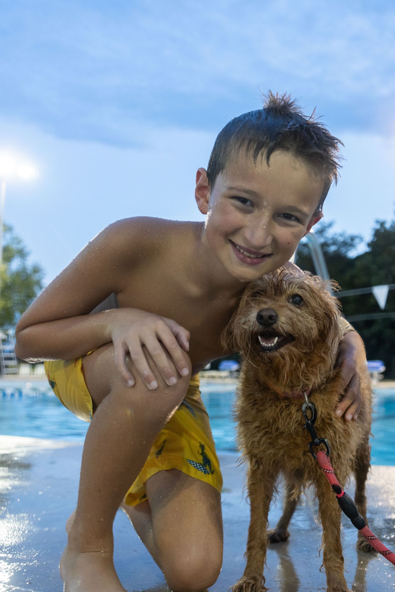 Smiling boy in yellow swim trunks hugging a wet brown dog by a pool at dusk.