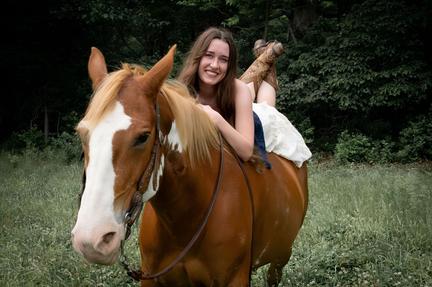 Smiling young woman with long brown hair riding a chestnut and white paint horse in a green field.