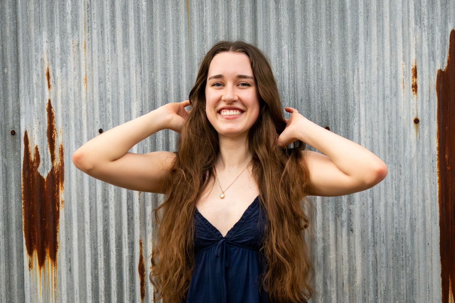 Smiling young woman with long wavy brown hair posing against a rustic corrugated metal wall.