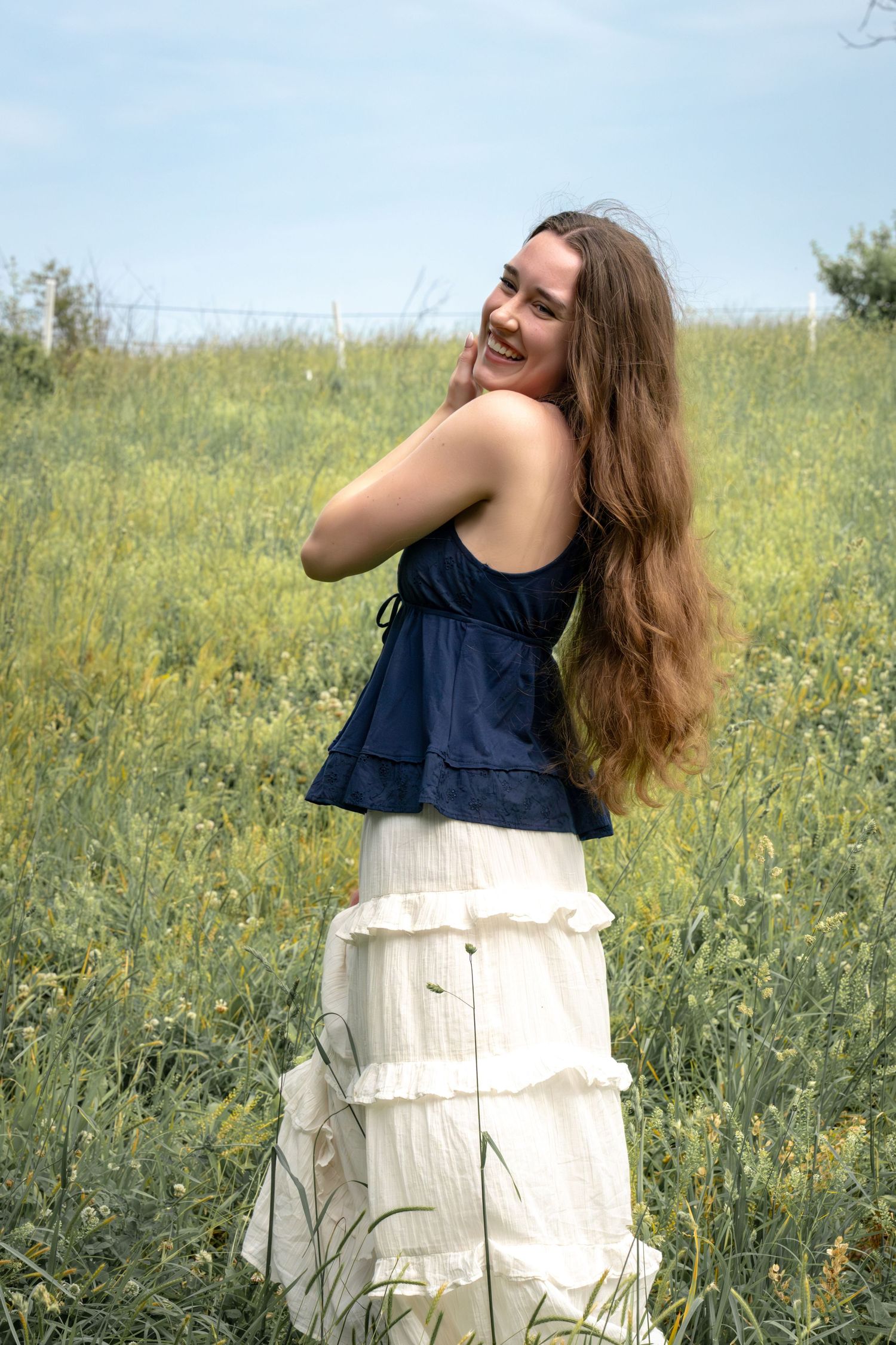 A smiling young woman with long brown hair wearing a navy top and white ruffled skirt standing in a green field.