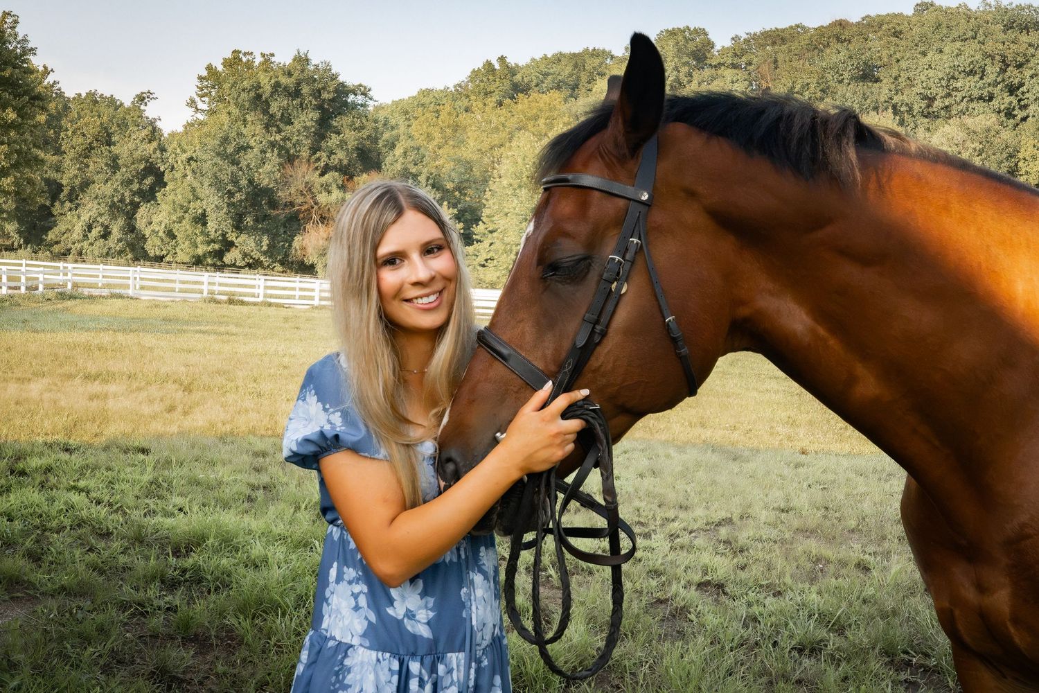 Young blonde woman in blue dress smiling beside a bay horse in a green pasture with white fence.