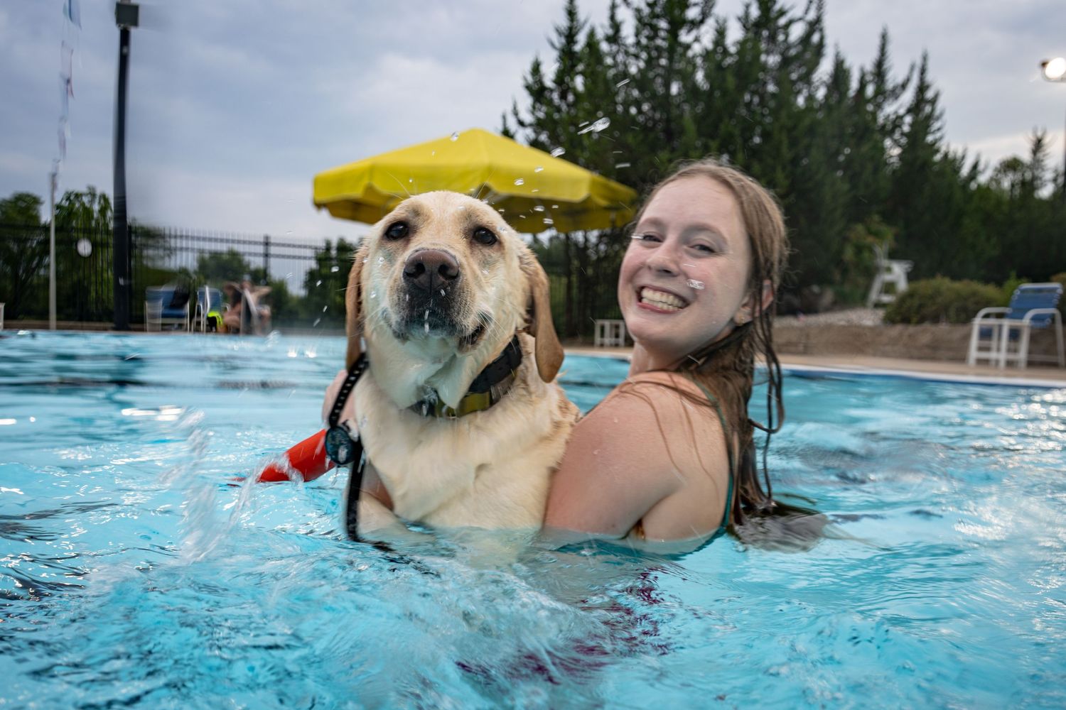 A smiling woman holds a yellow Labrador wearing a hat while swimming in a pool.