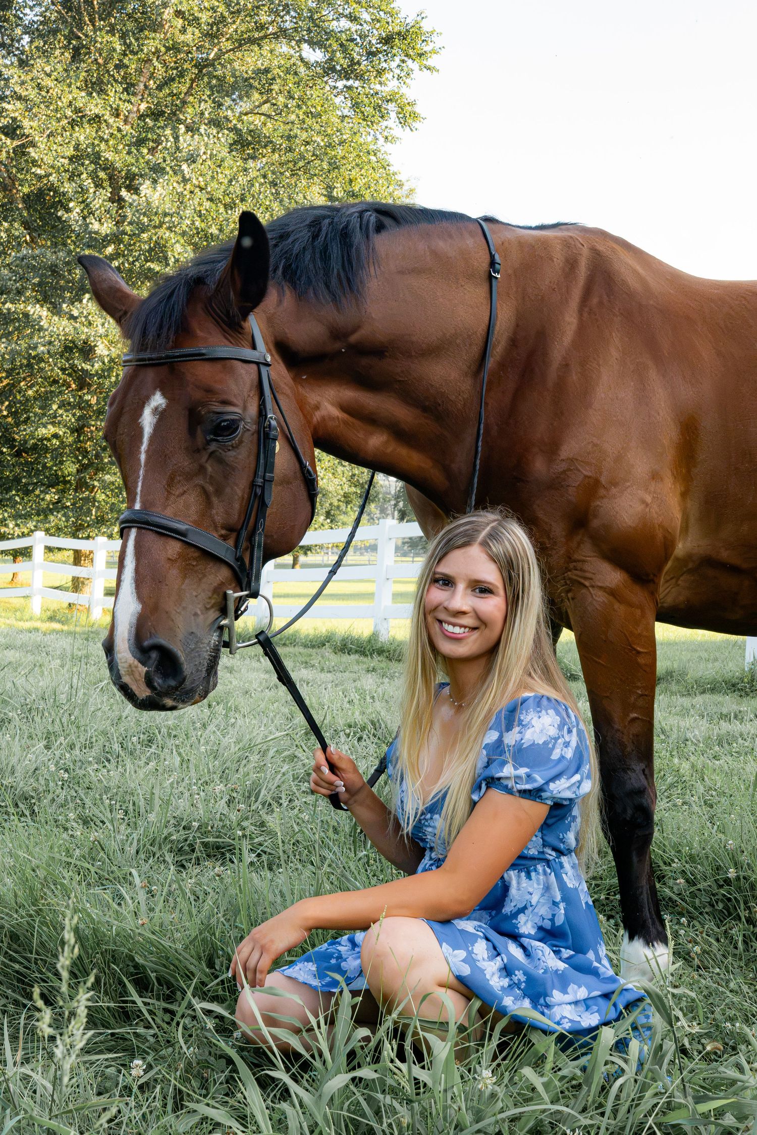 Young blonde woman in blue floral dress smiling beside a bay horse on green grass near white fence.