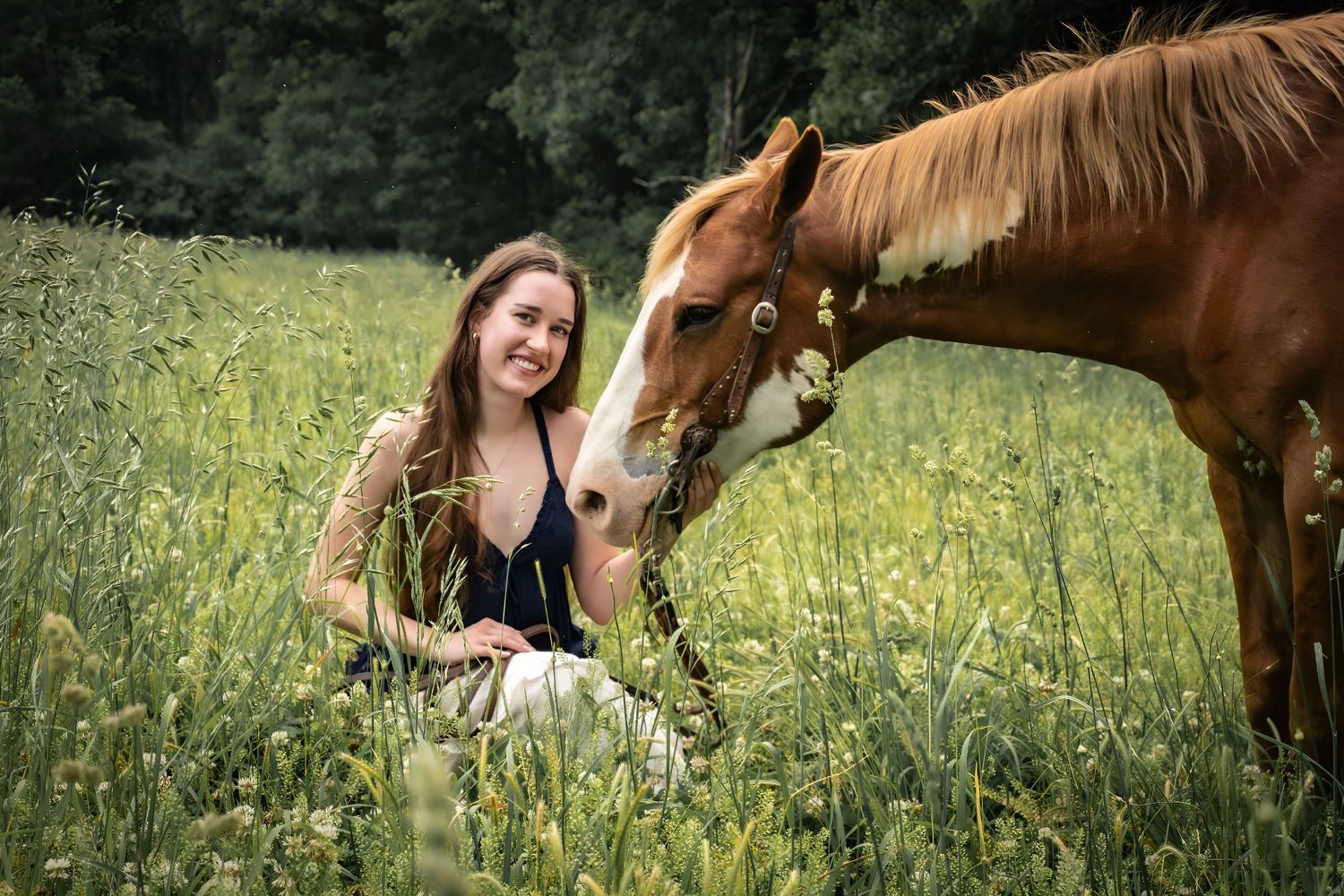 Young woman sitting in tall grass smiling as a chestnut horse nuzzles her in a green field.
