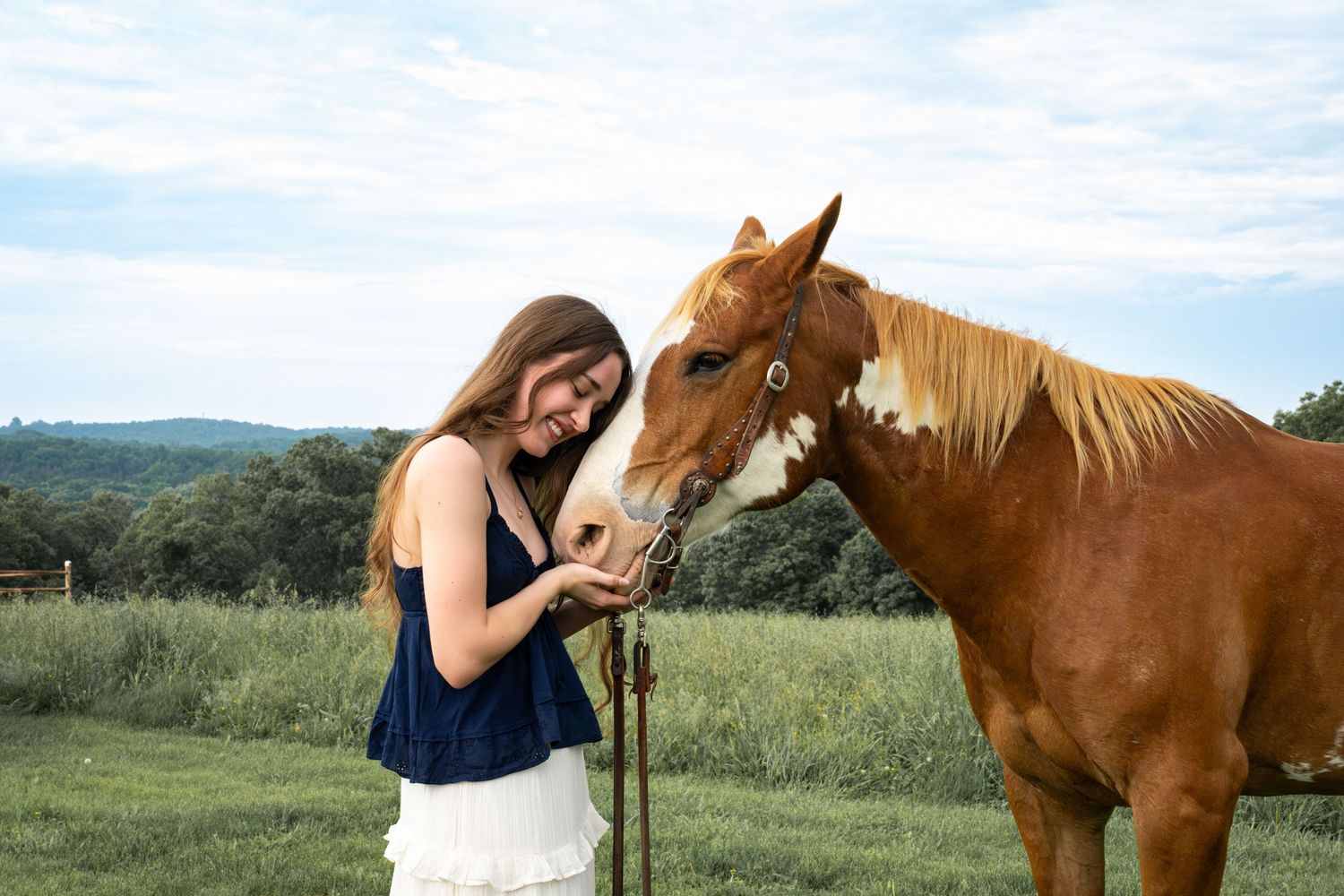 Young woman in navy top feeding or petting a chestnut horse with a white blaze in a green pasture.