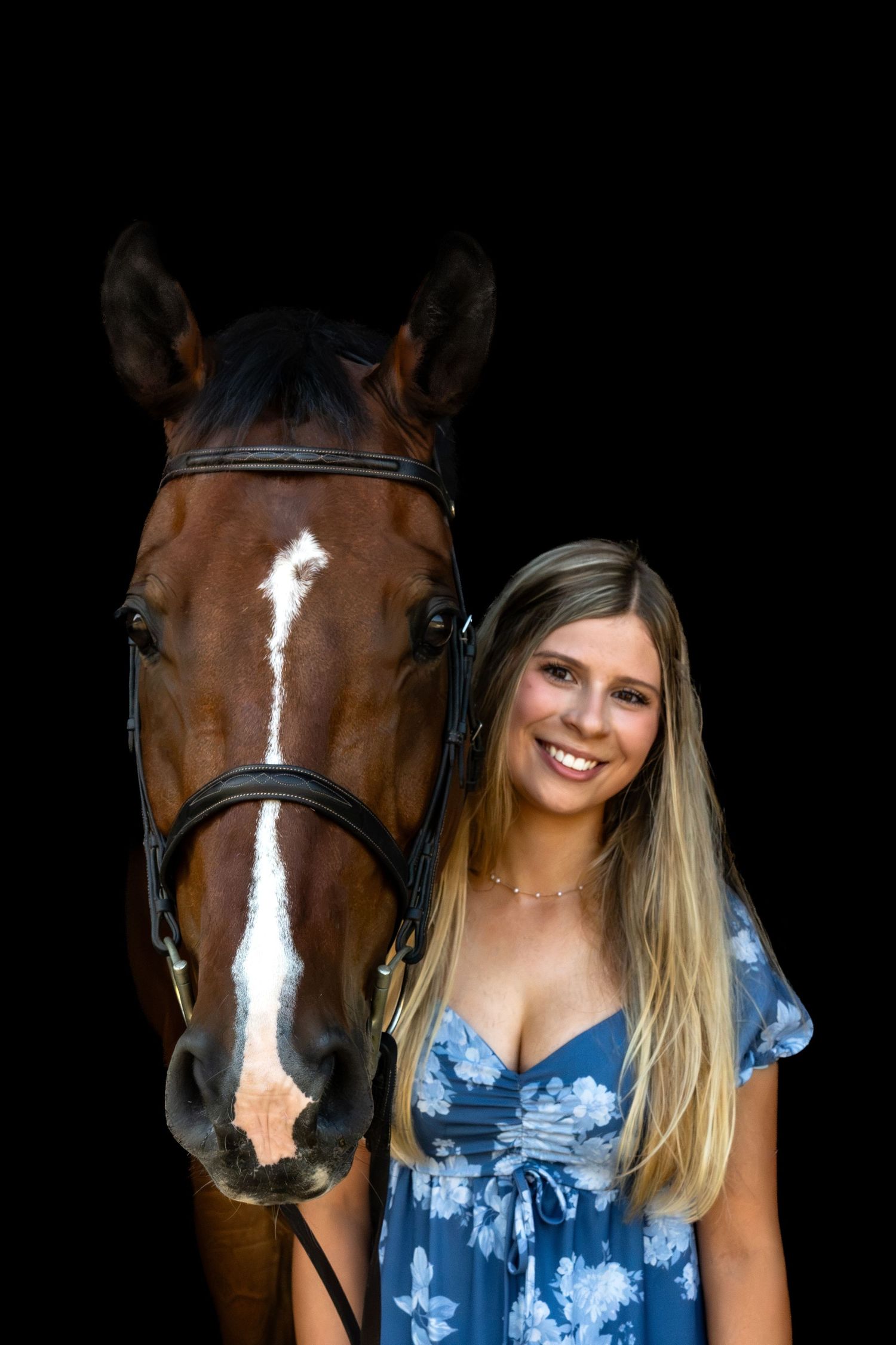 Smiling blonde woman in blue floral dress posing with a brown bay horse wearing a bridle against black background.