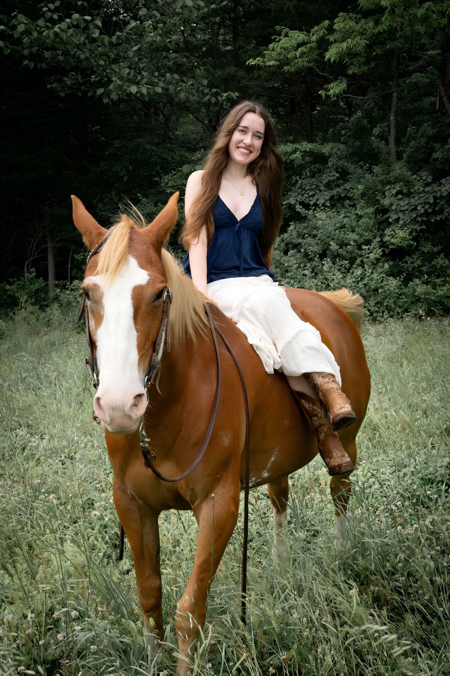 A smiling young woman in a blue top and white skirt riding a chestnut Paint horse in a green meadow.