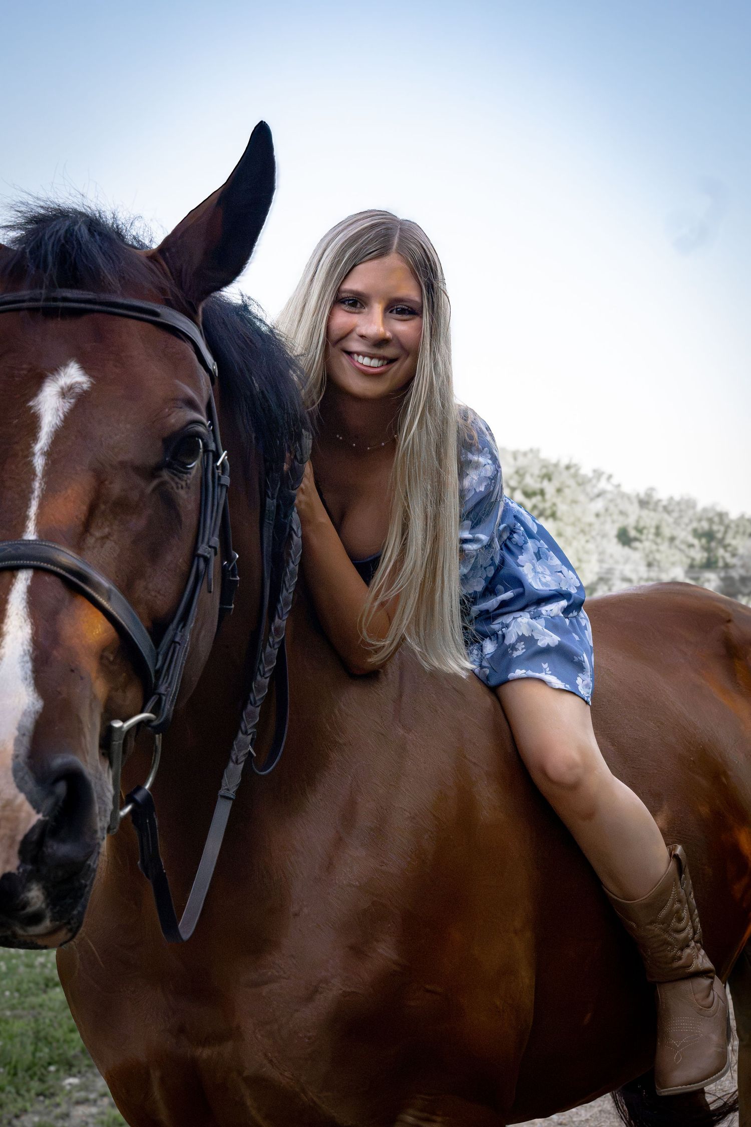 Smiling blonde woman in blue shirt riding a bay horse with a white blaze, outdoors with trees in background.